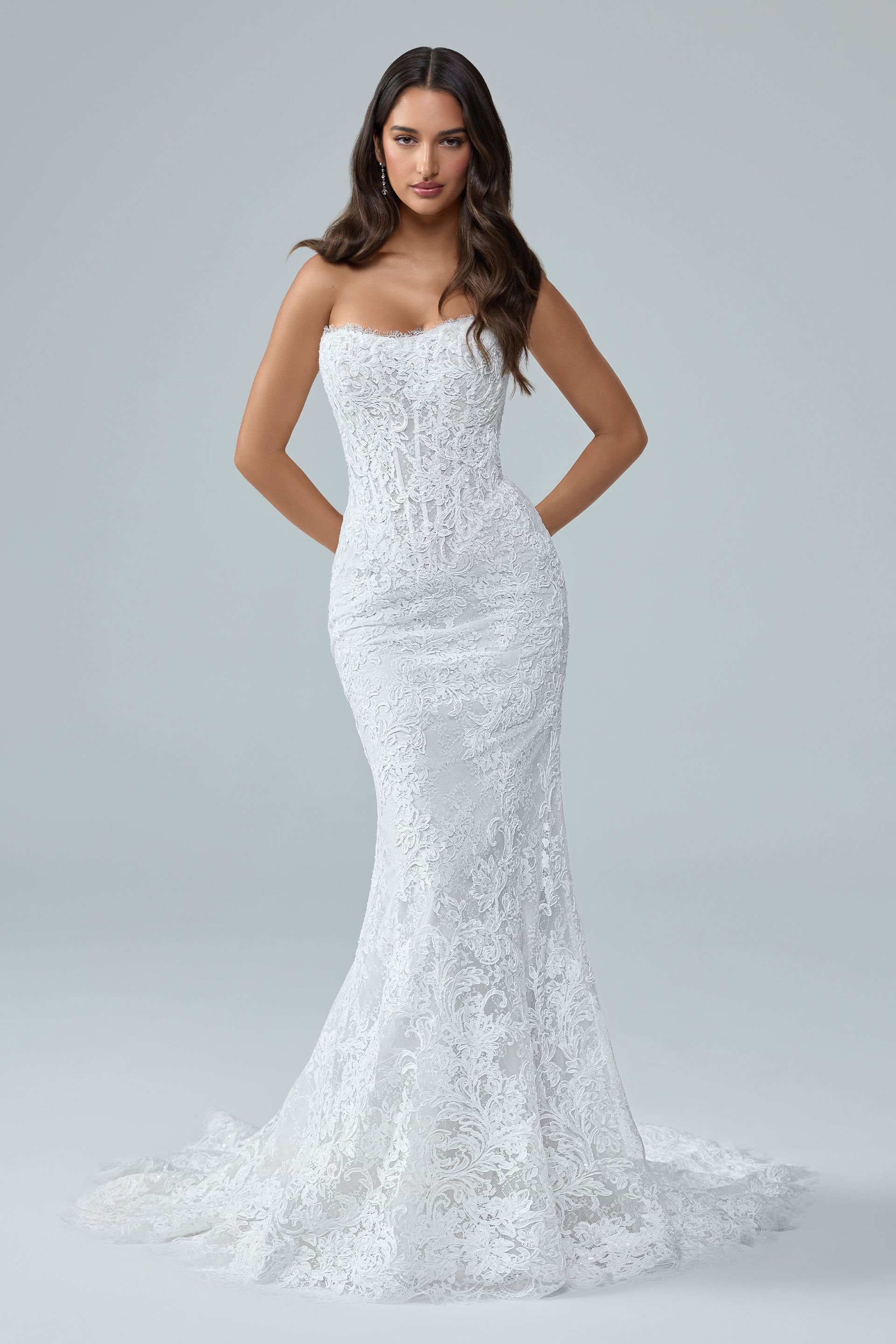 Woman in a strapless white lace mermaid gown standing against a gray background. Woman in a strapless white lace mermaid gown standing against a gray background.