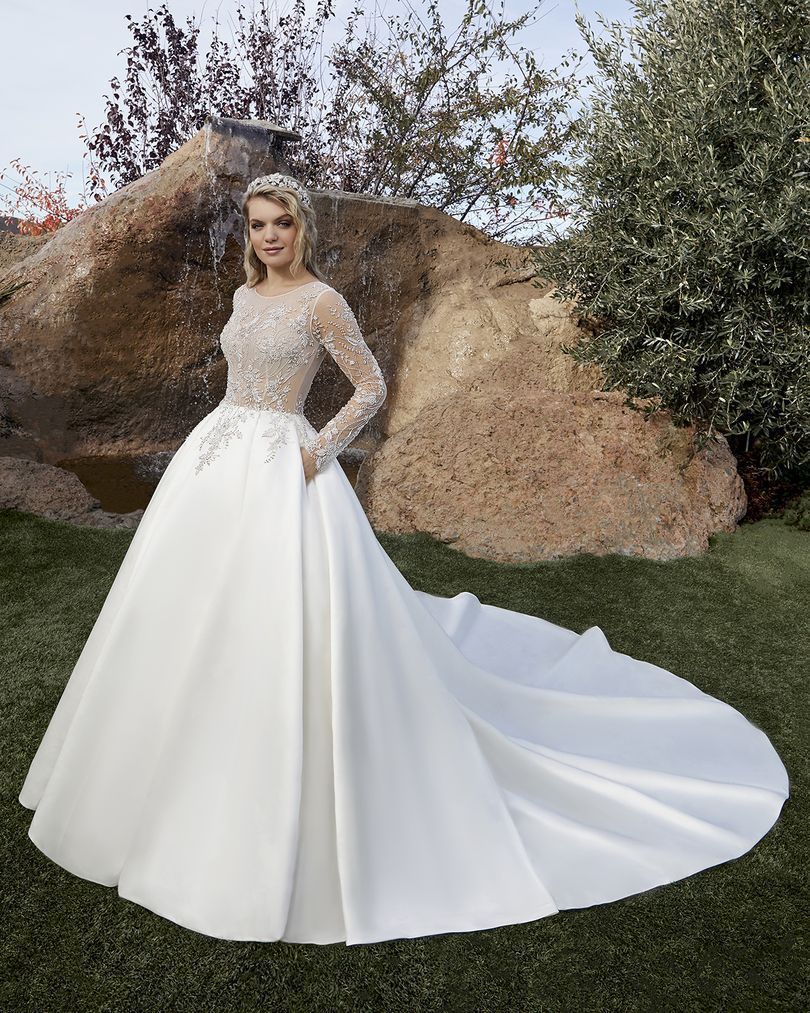 Woman in a long white satin wedding gown with a beaded bodice and long sleeves, outdoors near a rock.