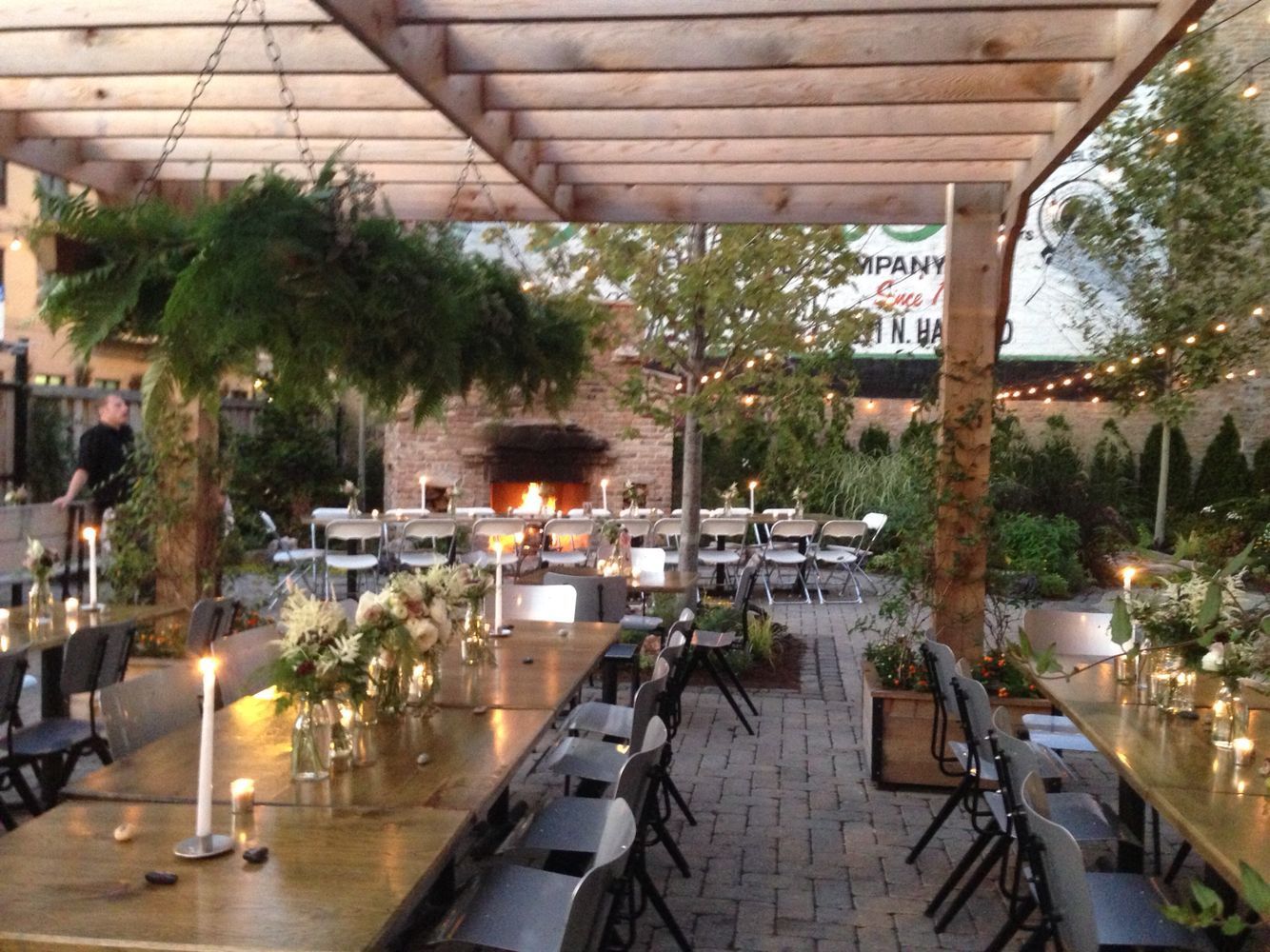 Outdoor dining area with tables, chairs, fireplace, overhead pergola, and string lights, set for a formal event.