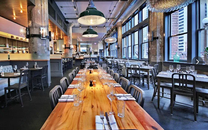 Restaurant interior with long wooden tables set for diners under industrial-style lighting.