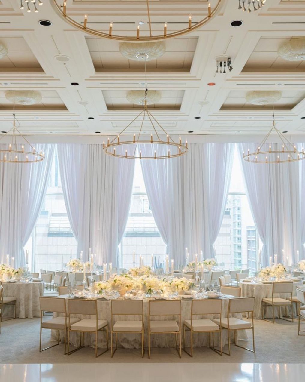 Elegant ballroom set for a wedding, with white drapes, gold chandeliers, and tables set for guests.