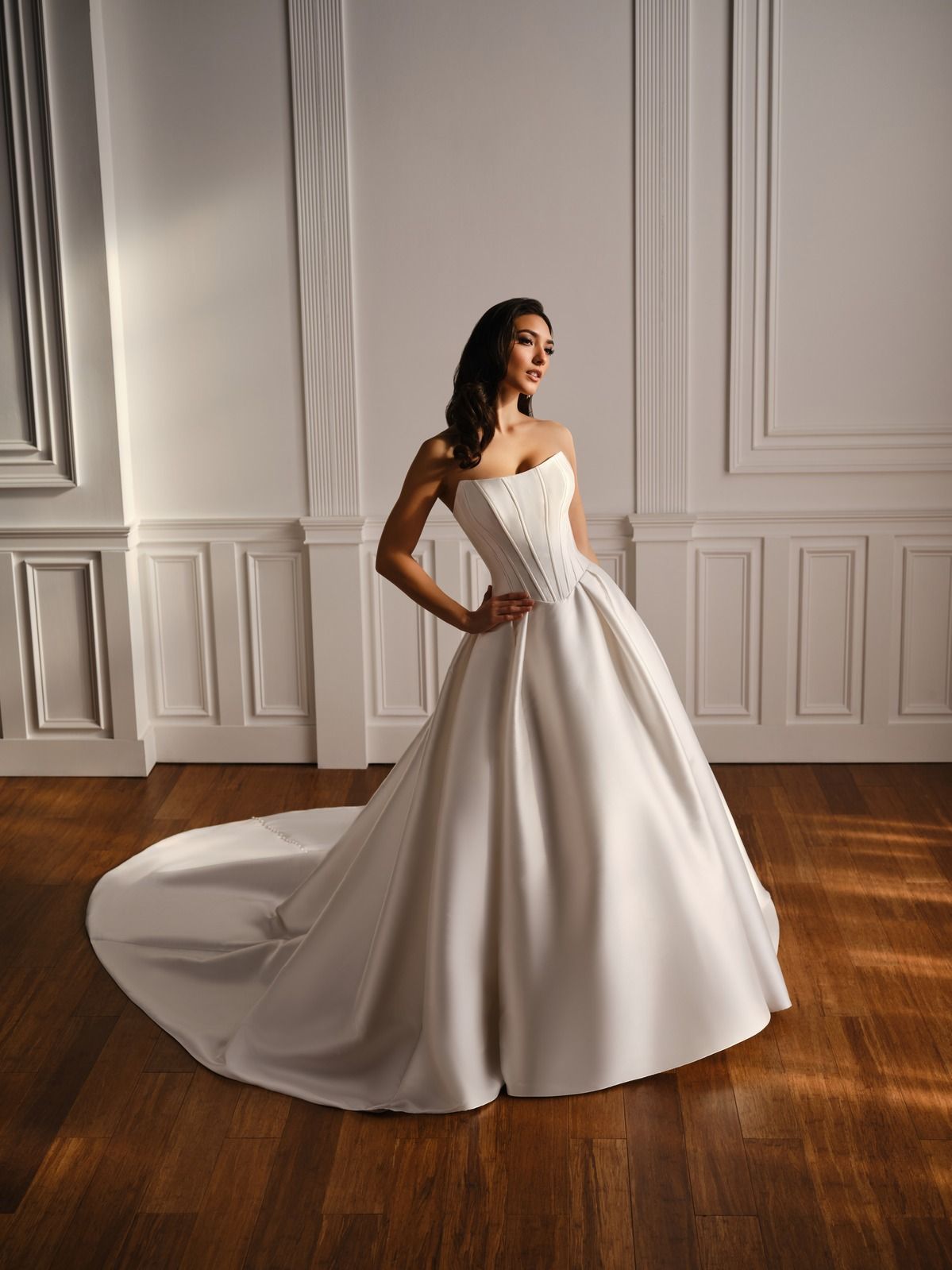 Woman in a white strapless gown posing in a bright room with paneled walls and a wood floor.