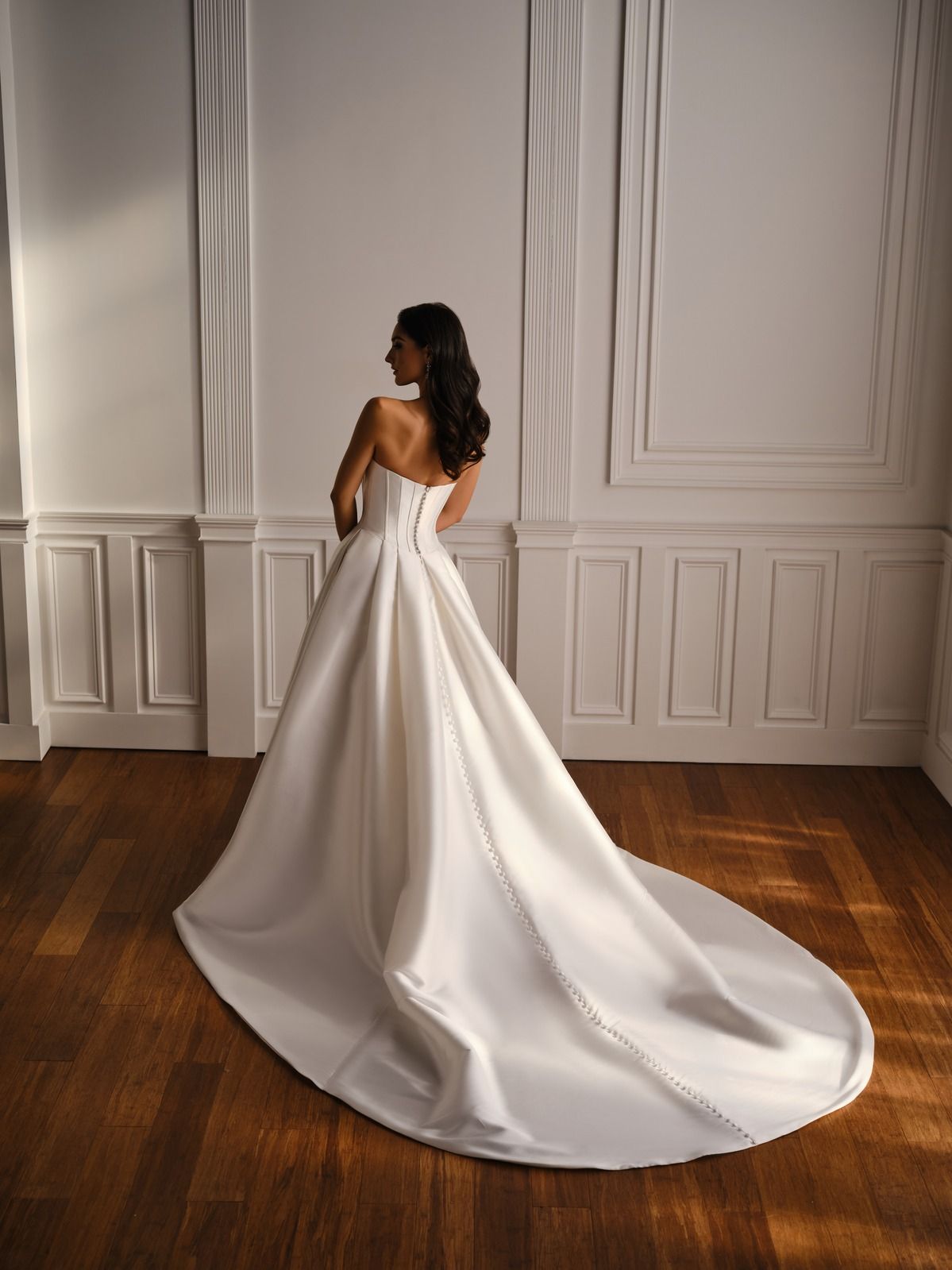 Woman in a white strapless gown posing in a bright room with paneled walls and a wood floor.