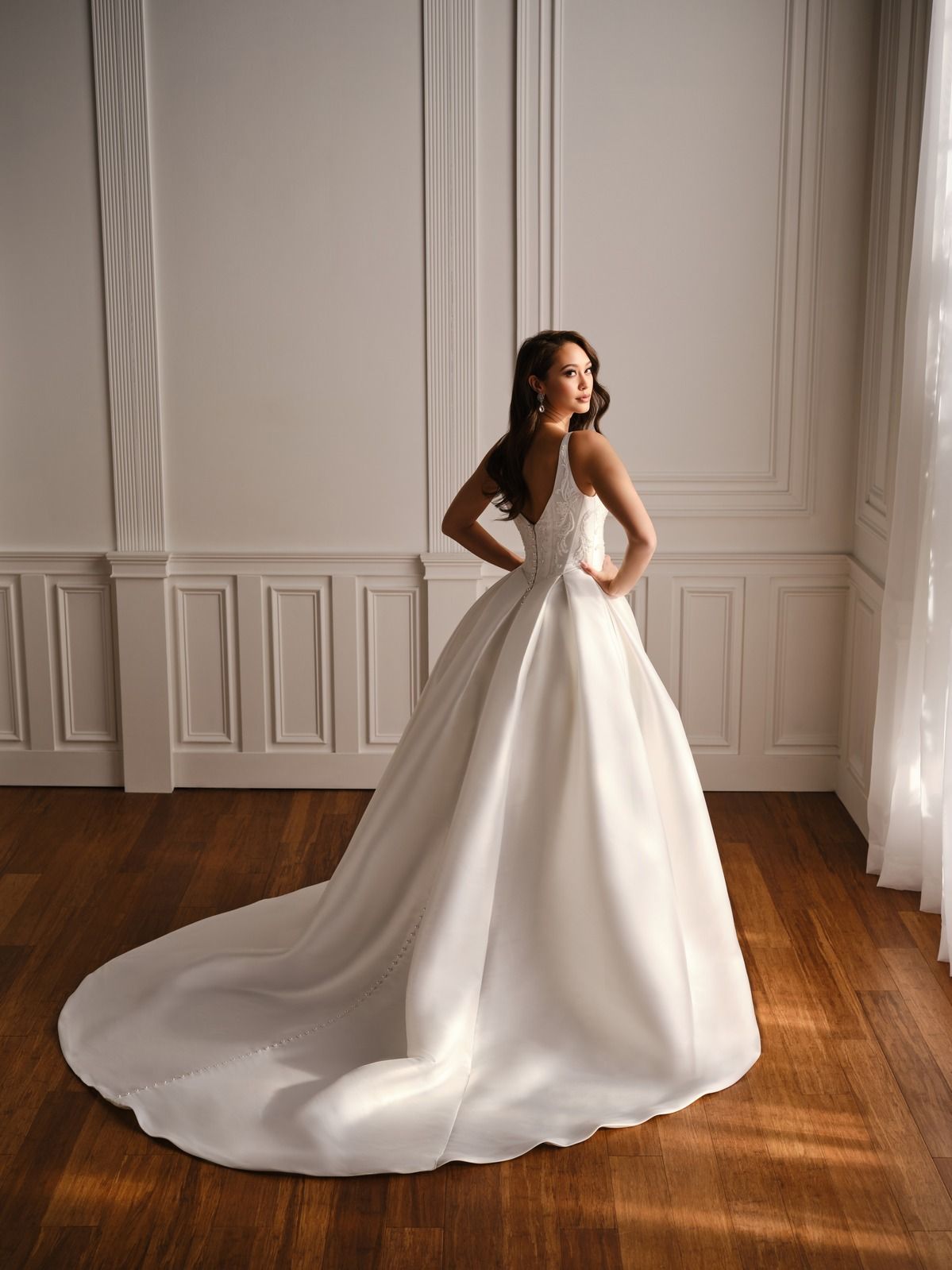 Woman in a white bridal gown posing in a bright room with wooden floors and sheer curtains