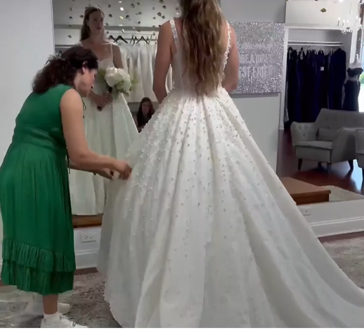 Woman in green dress adjusting a bride's white wedding gown in a boutique. Mirror reflection and decor visible.