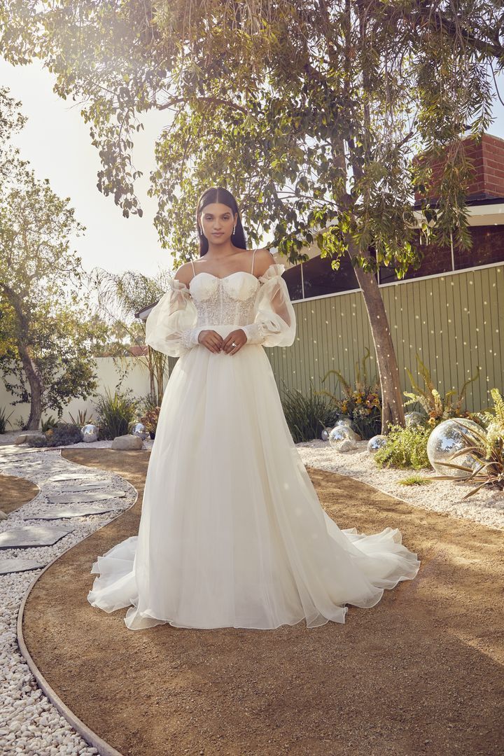 Woman in a white off-shoulder wedding dress stands outdoors, hands clasped.