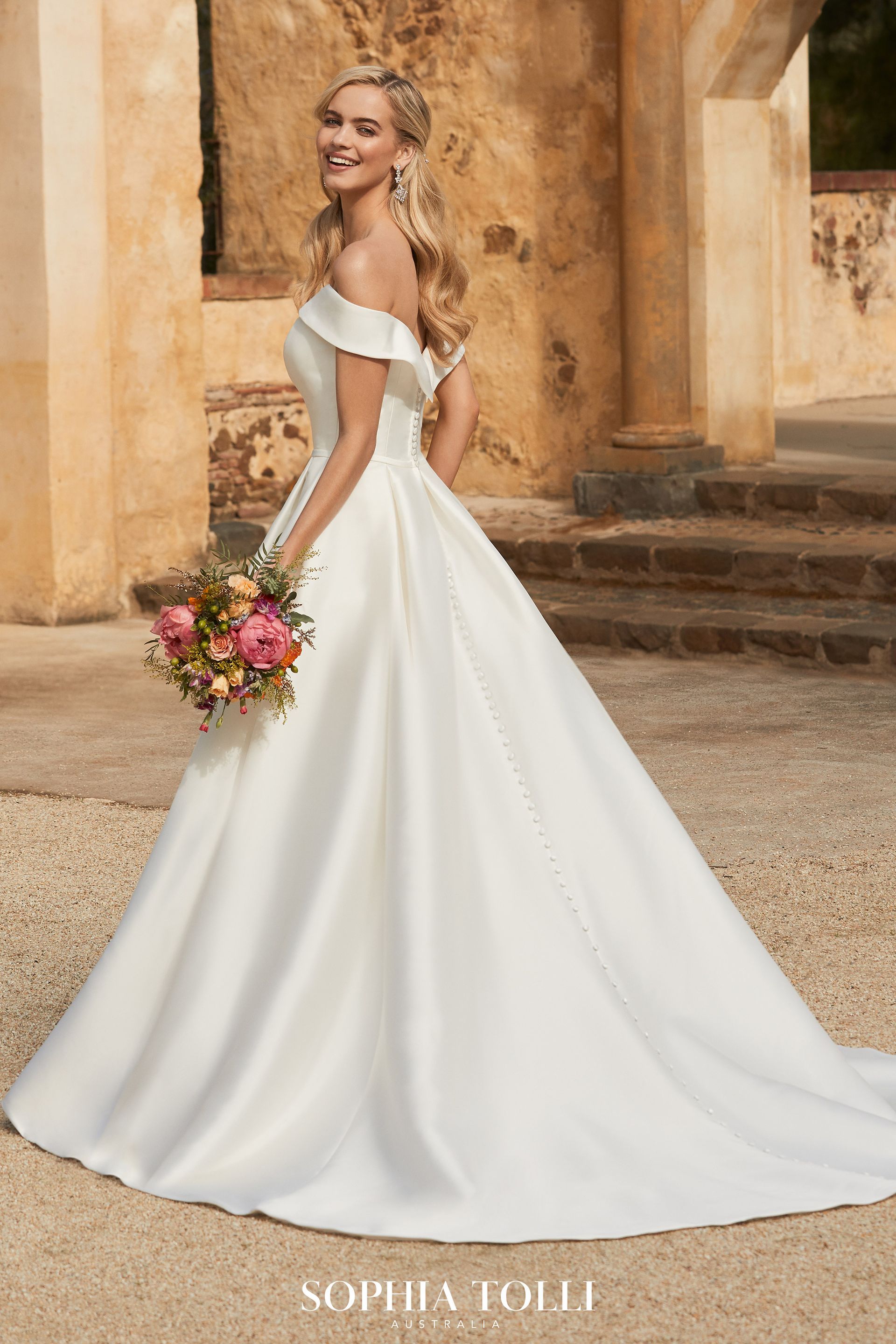 Woman in white wedding dress, holding flowers, smiling, standing outside a building.