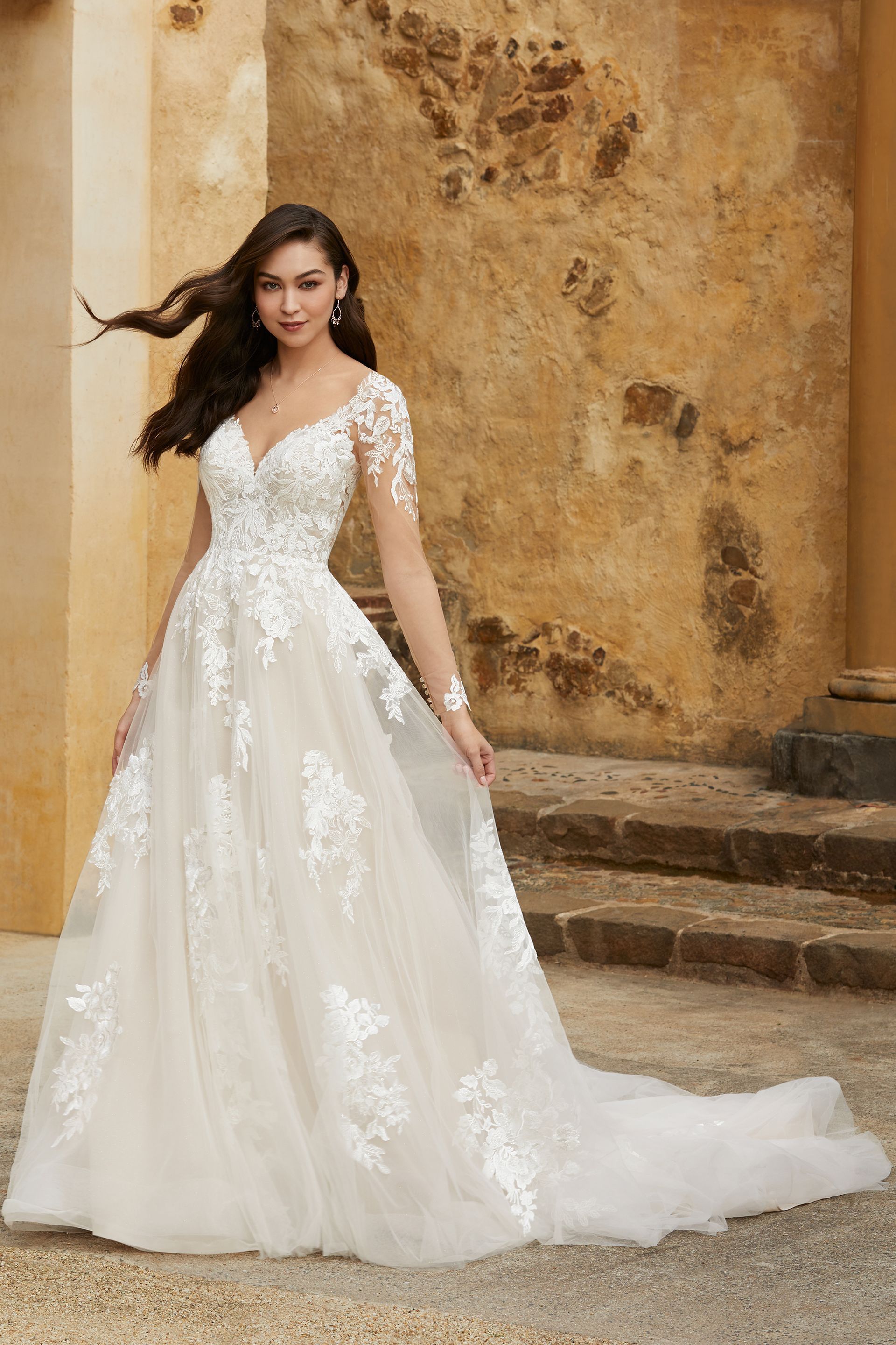 Woman in a white wedding gown with lace detail, standing outdoors with a stone wall backdrop.