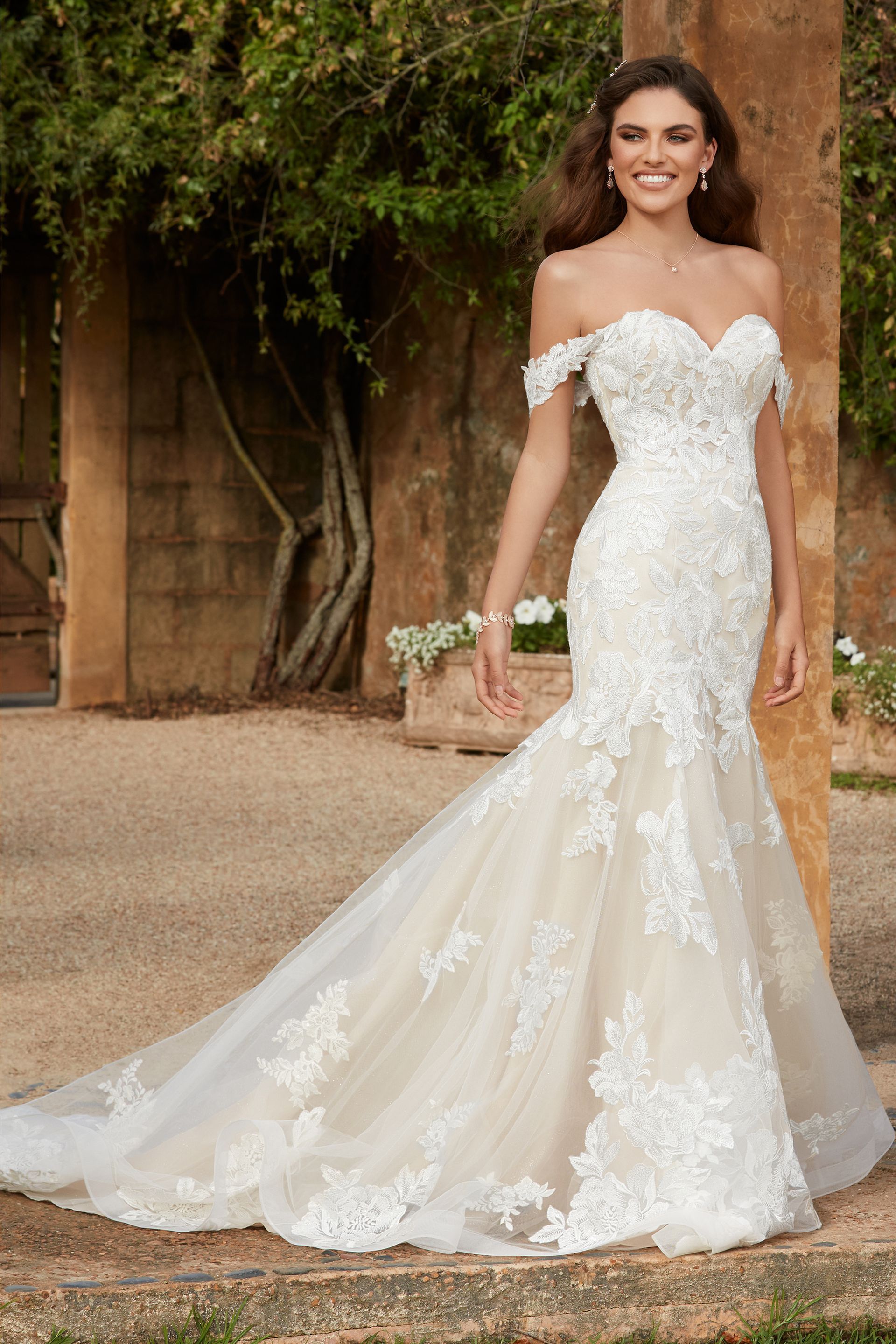 Woman in a white off-the-shoulder lace wedding dress, smiling, outdoors.