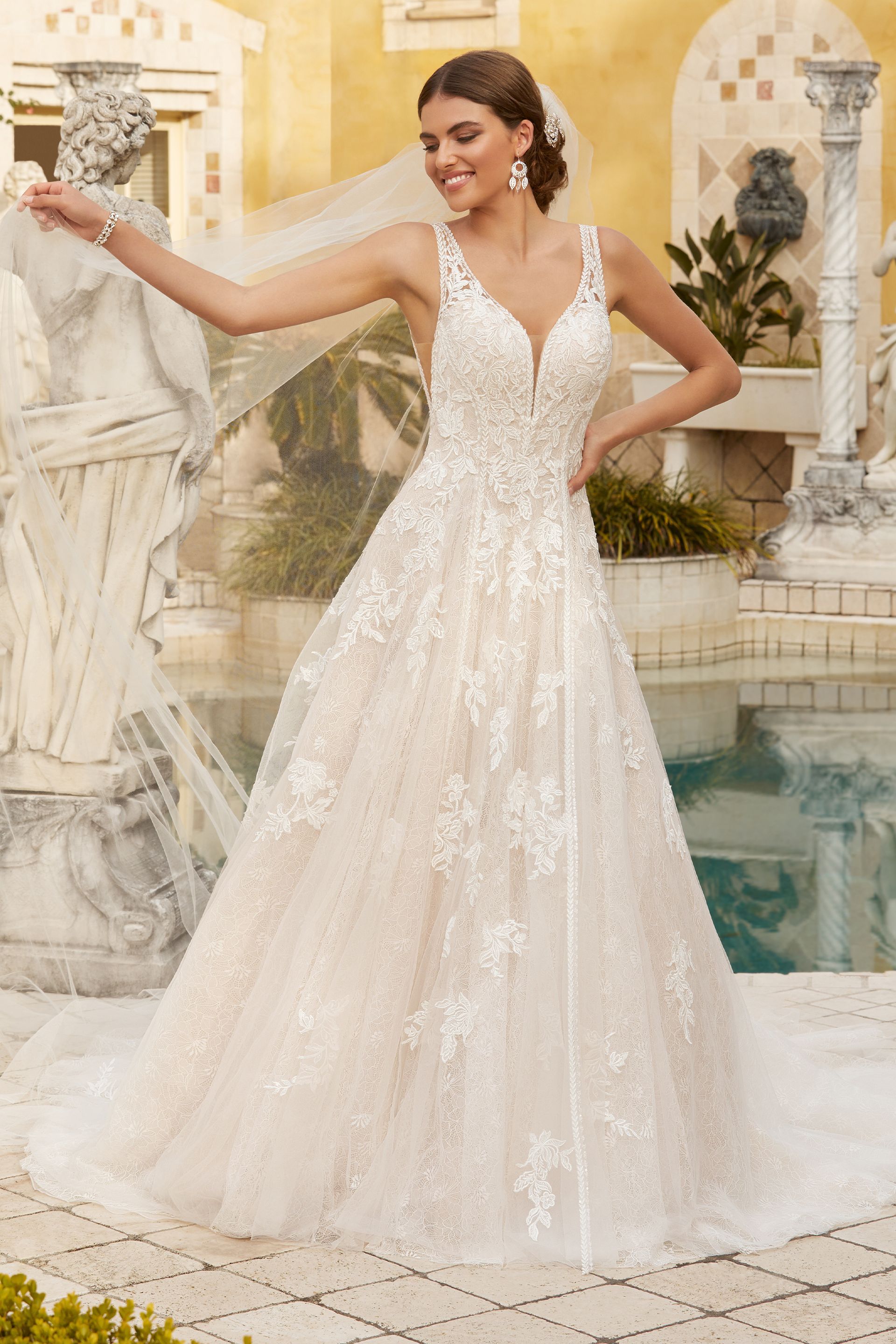 Woman in a wedding dress, posing outdoors near a fountain. She is smiling, with floral lace details.