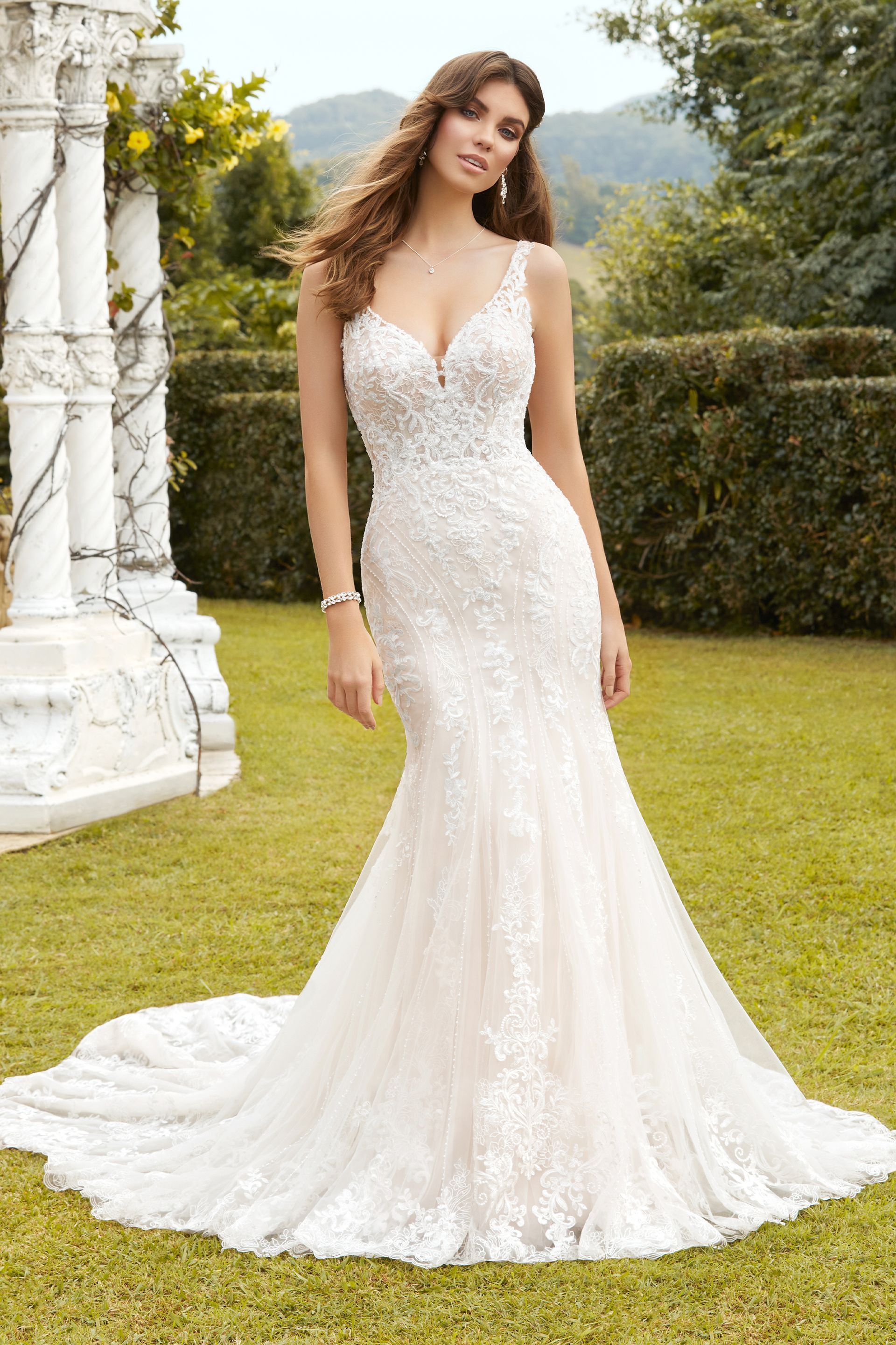 Woman in a white lace mermaid wedding dress, standing outdoors in a garden setting.
