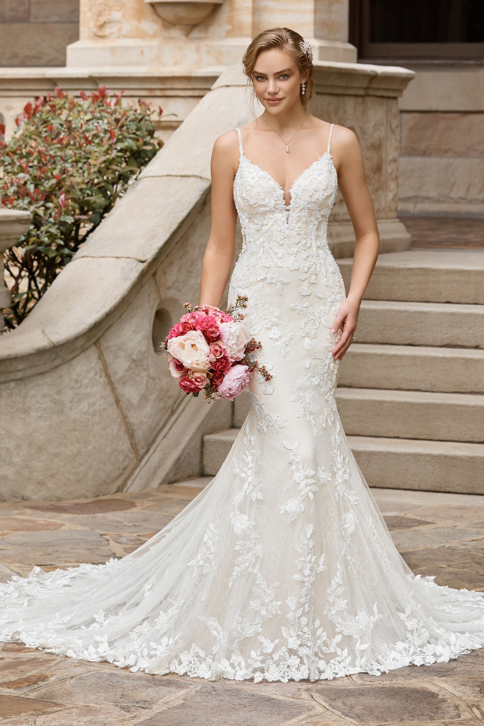 Woman in white lace wedding dress, holding bouquet, standing on stone steps.