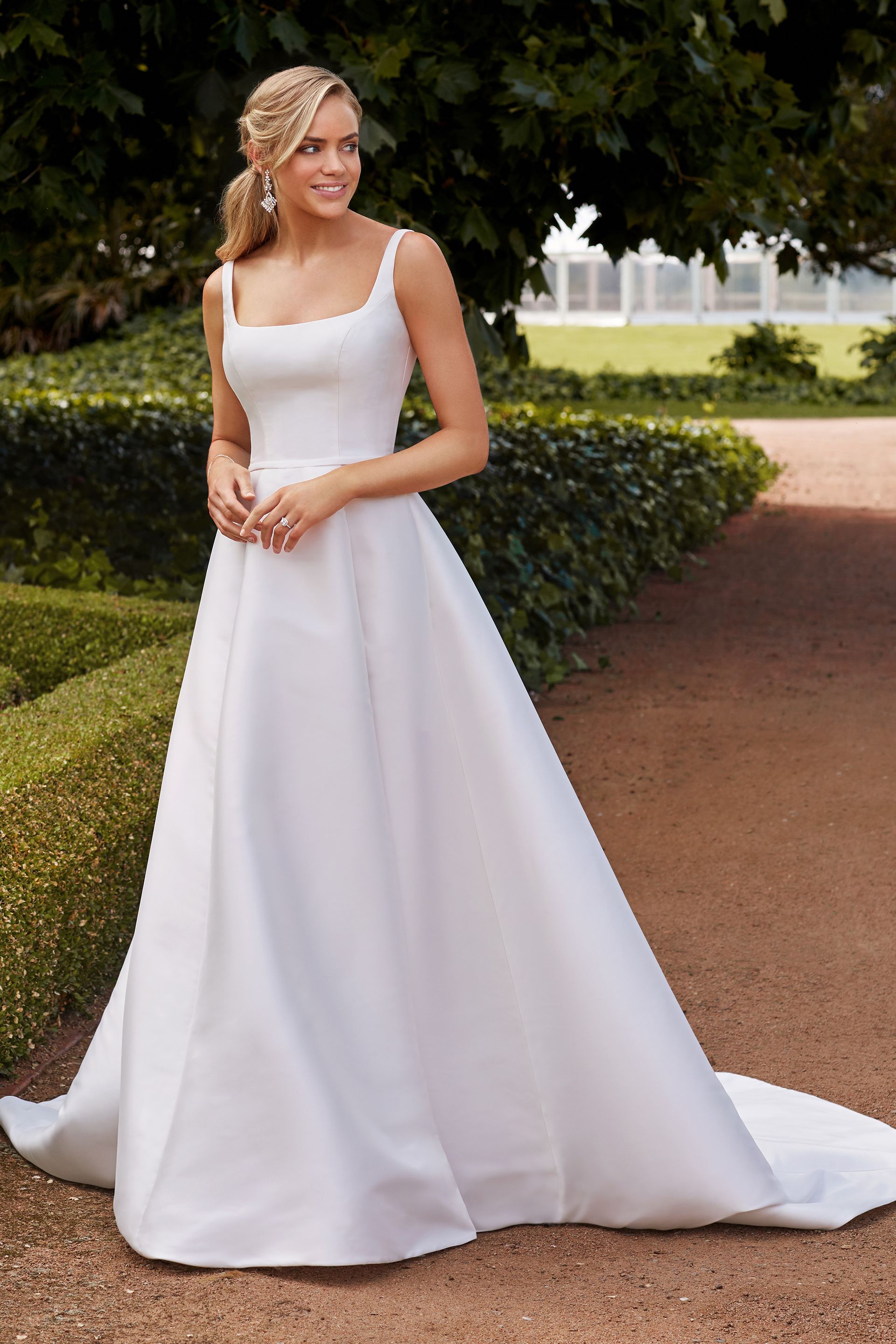 Woman in a white wedding dress smiles, standing outdoors on a pathway lined with greenery.