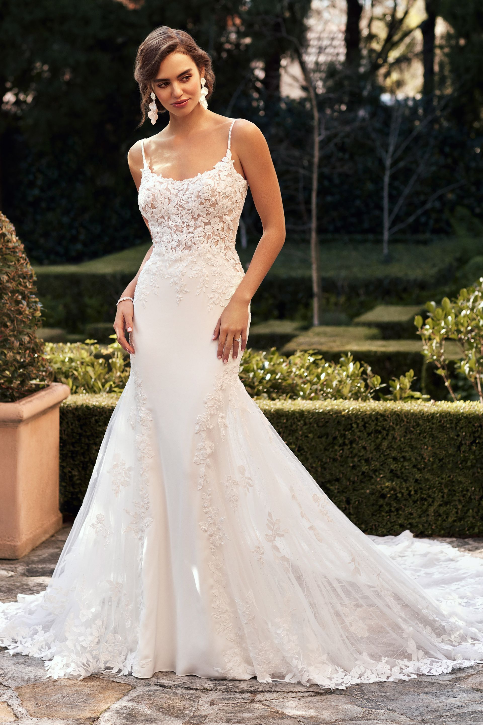 Woman in a white lace wedding dress with spaghetti straps and a train, standing outdoors.
