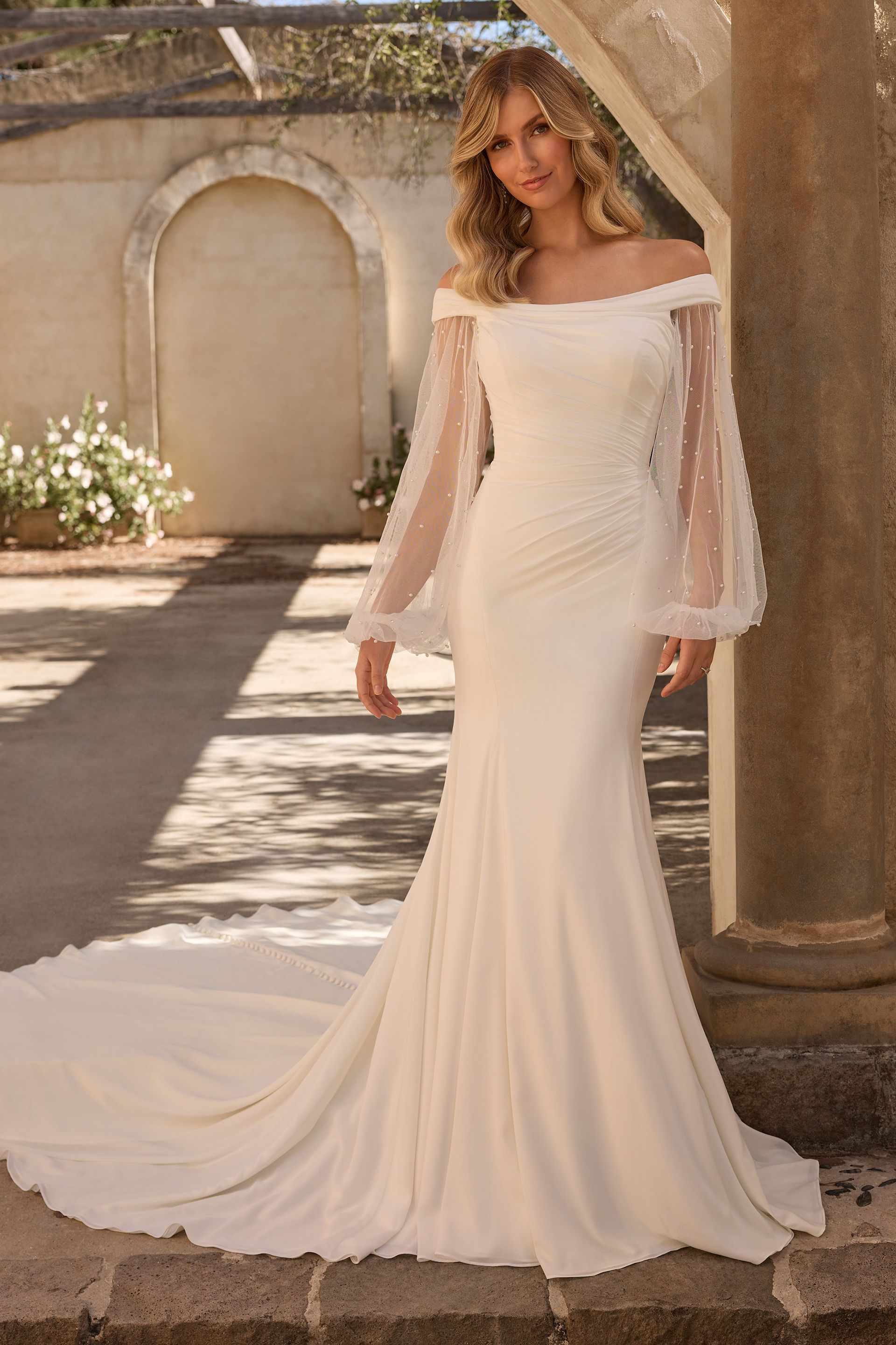 Woman in an ivory off-shoulder wedding gown standing in a sunlit courtyard with a long train Woman in an ivory off-shoulder wedding gown standing in a sunlit courtyard with a long train