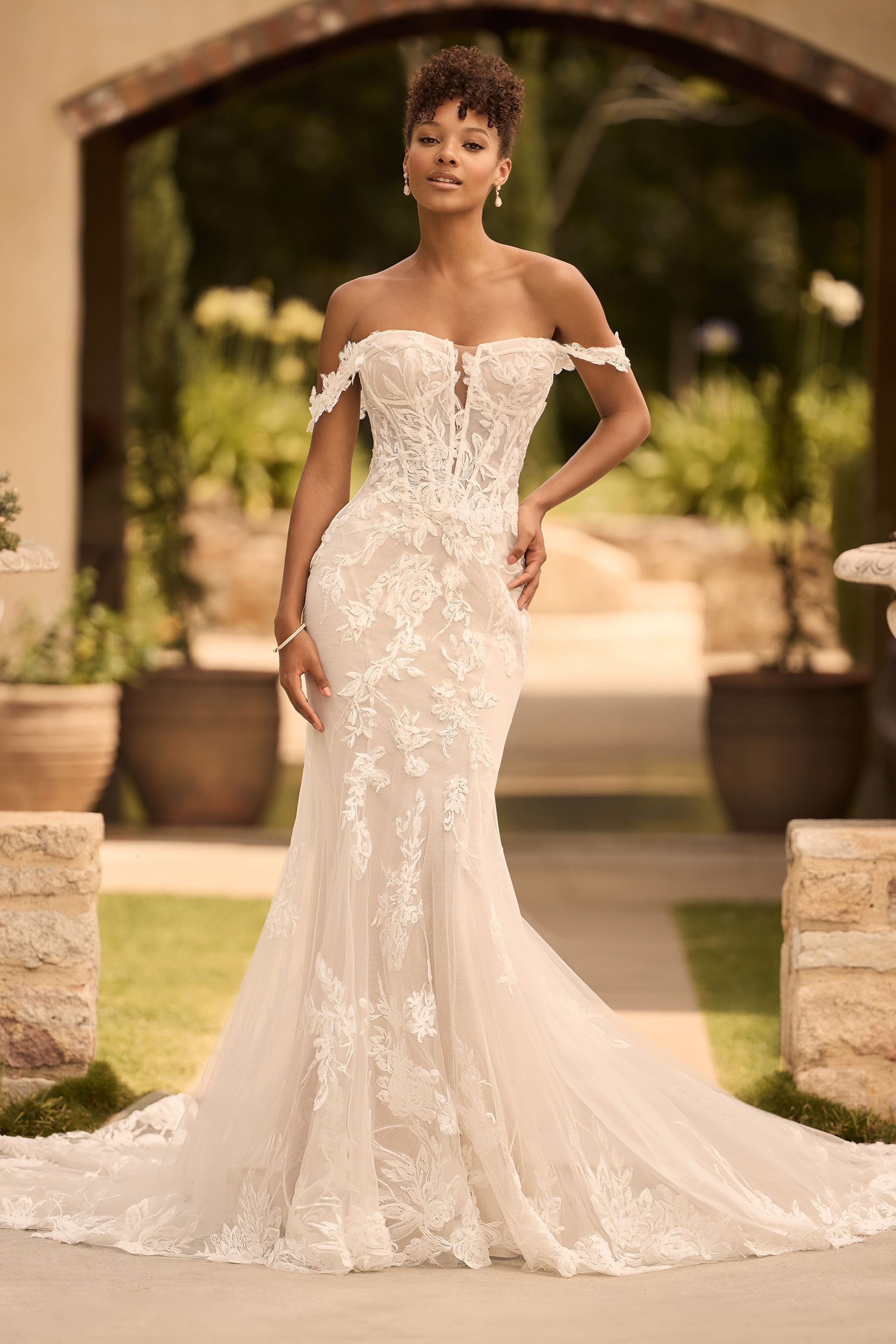 Woman in a white, off-the-shoulder wedding gown with lace detailing, outdoors in a garden setting.