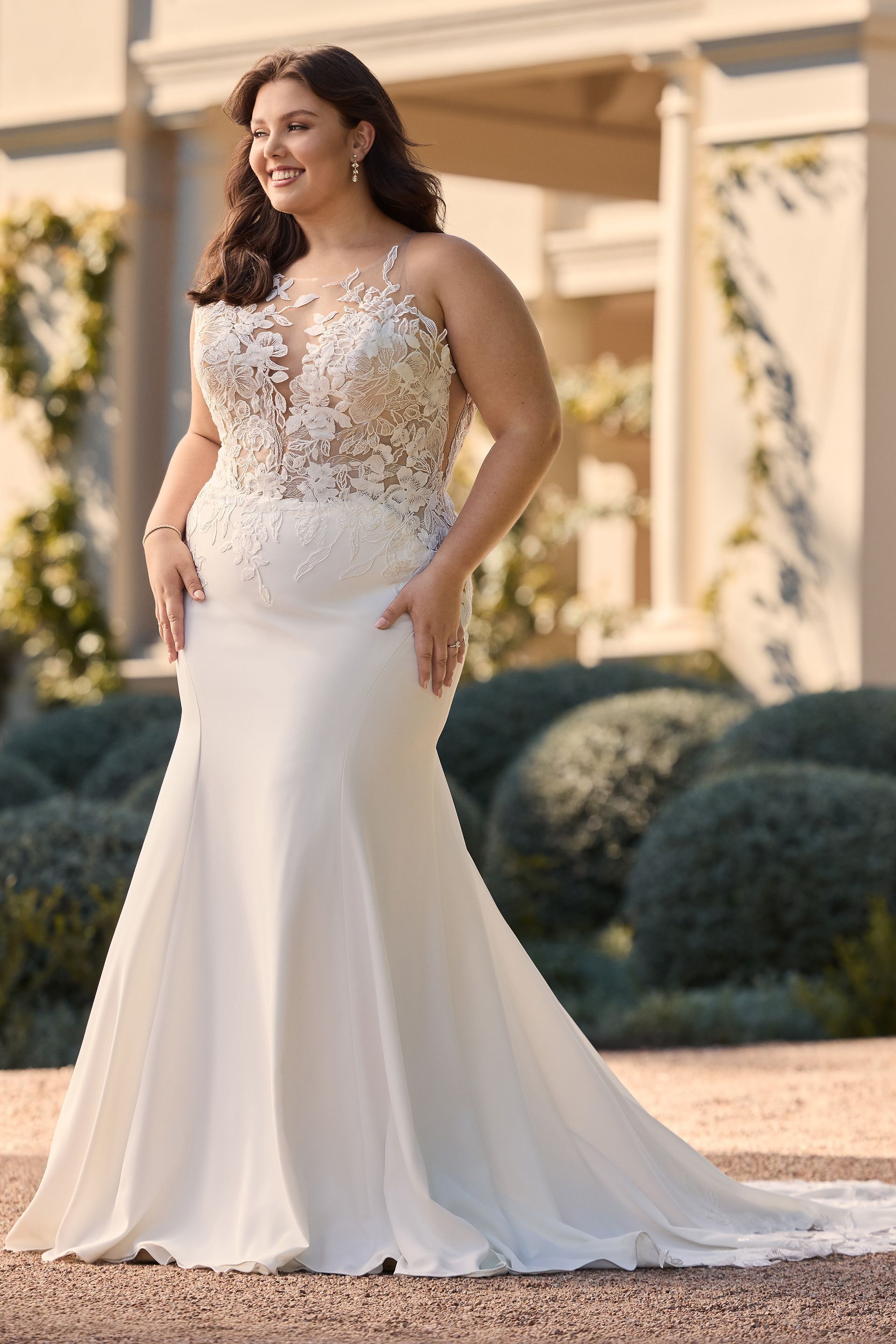 Woman in a white wedding dress, smiling. She is outdoors with a garden backdrop.