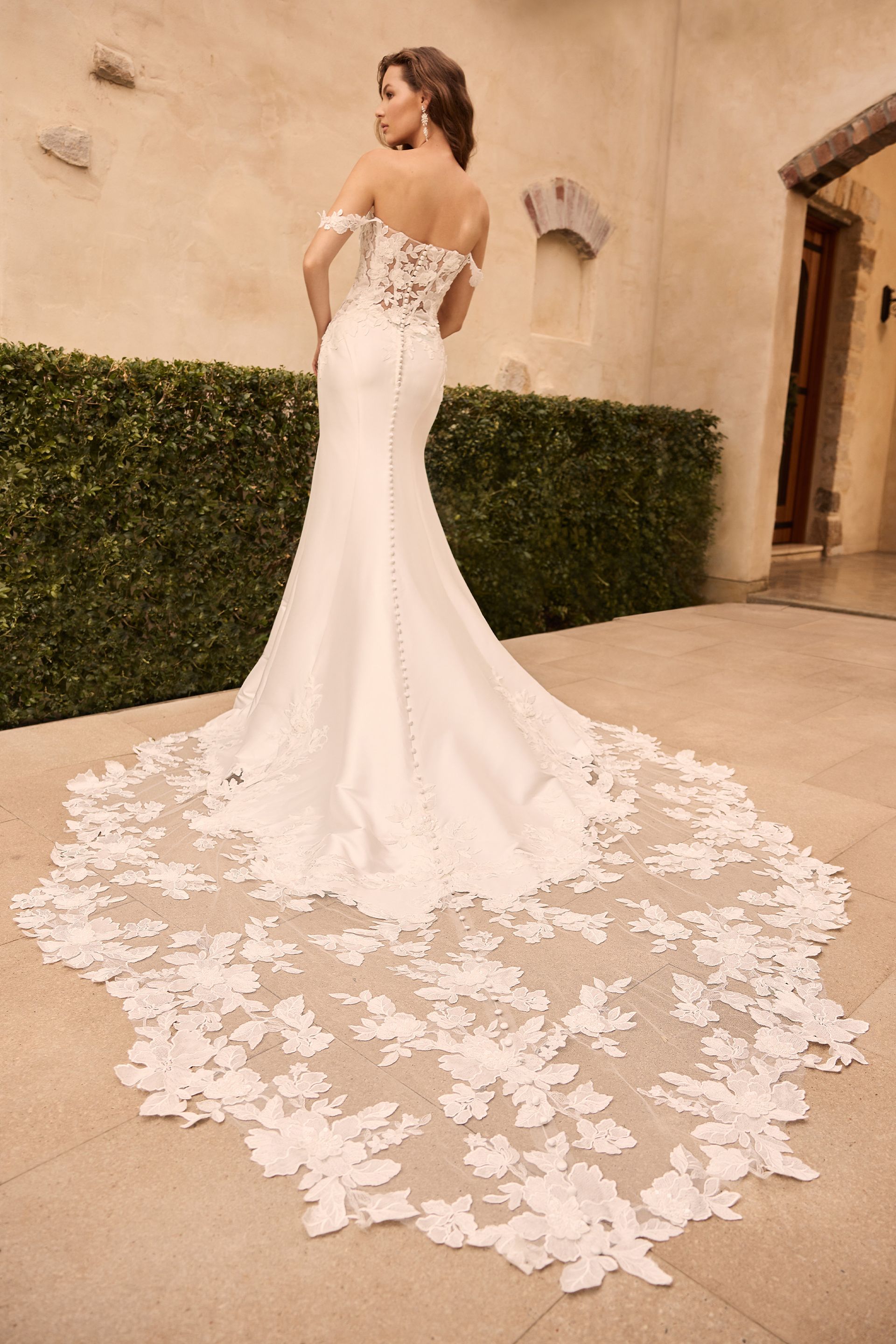 Woman in off-the-shoulder wedding dress with lace bodice, long train, posing outdoors in front of a stone building.