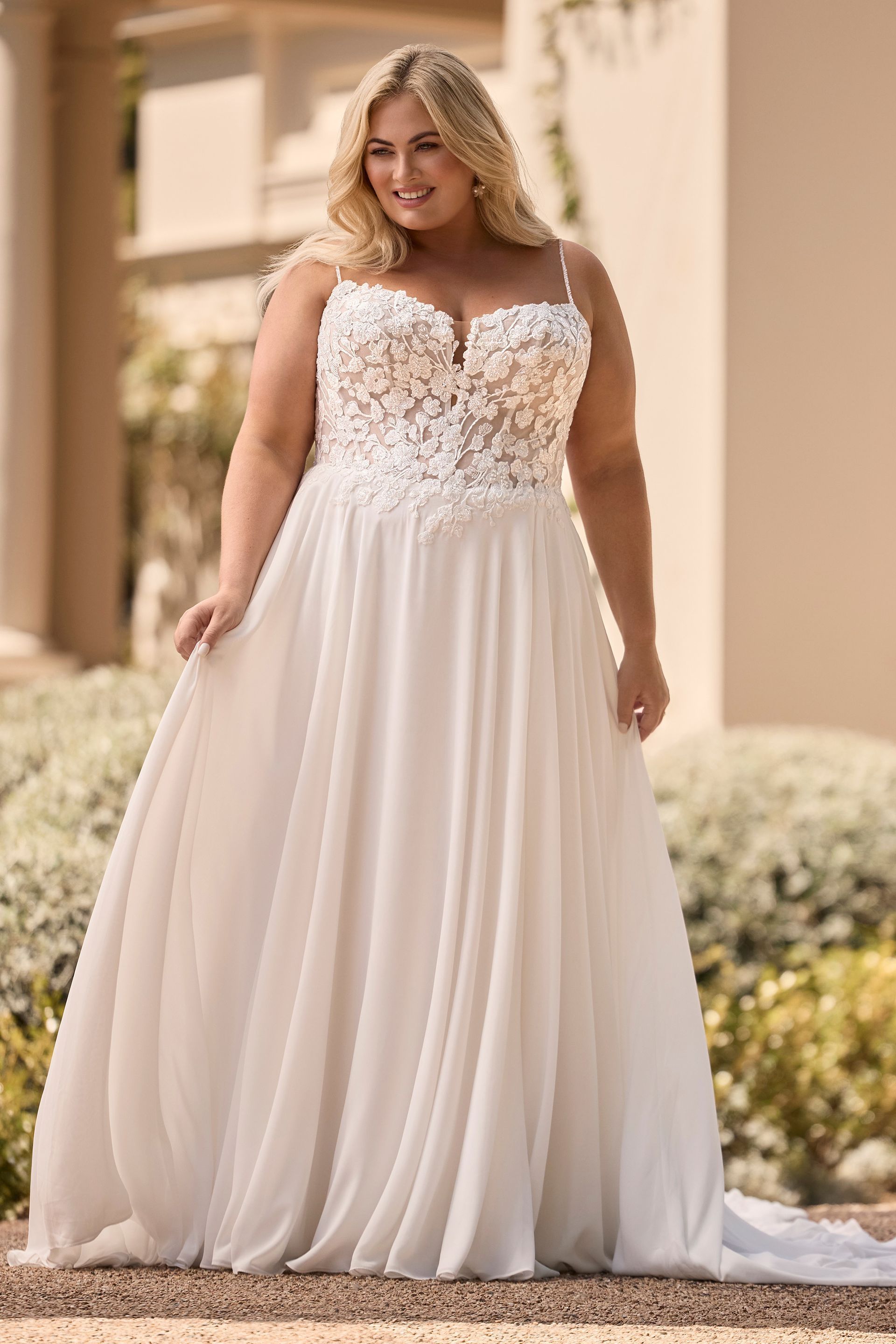 Woman in white wedding dress, floral bodice, spaghetti straps, standing outdoors, smiling.
