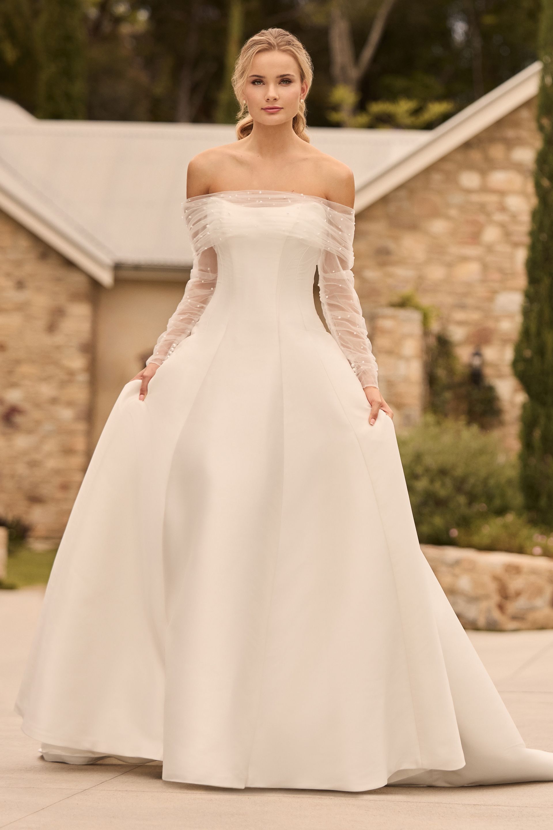 Woman in off-the-shoulder wedding gown, holding out skirt, standing outdoors.