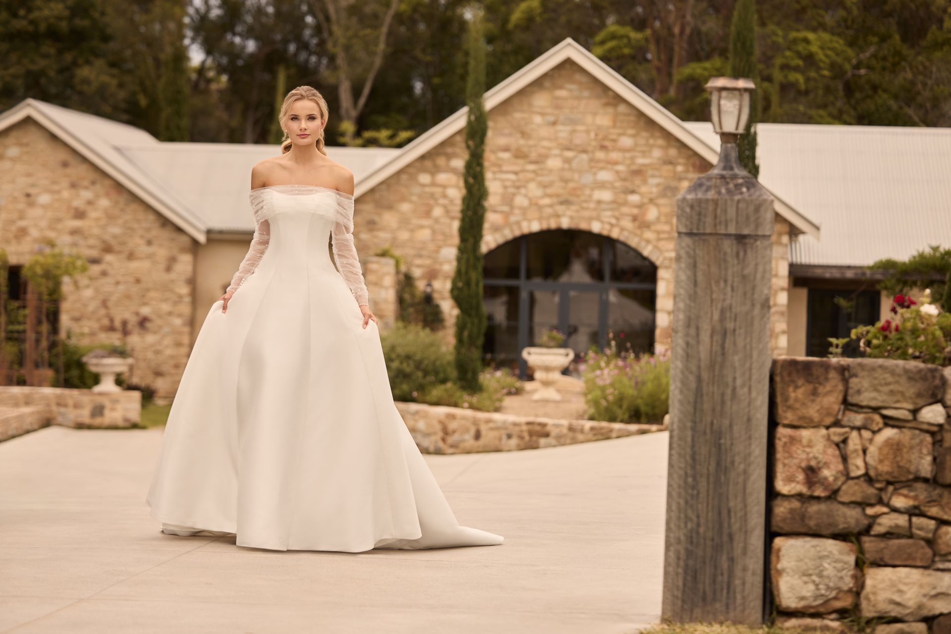 Woman in a white off-the-shoulder wedding gown stands in front of a stone building with arched windows.