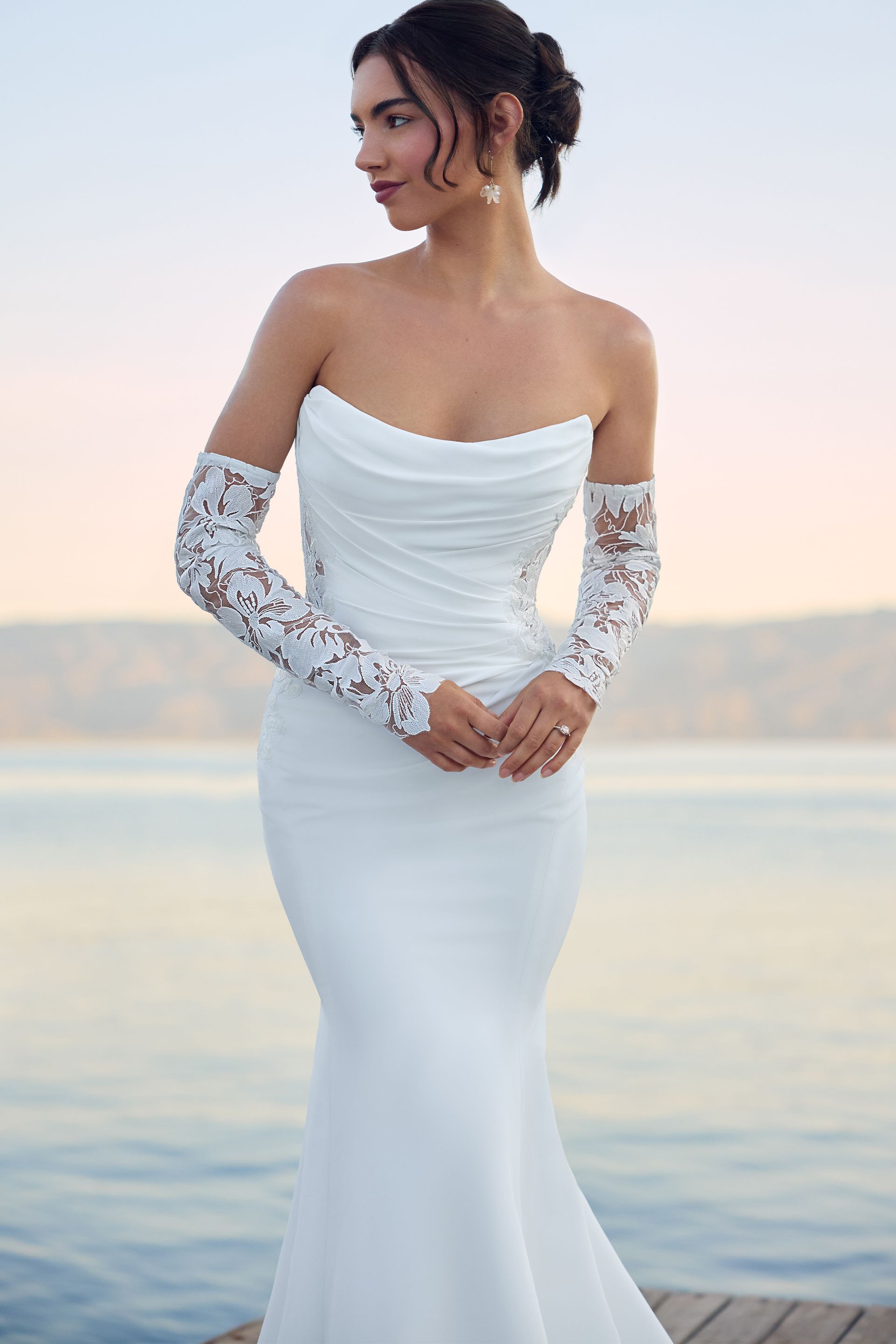 Woman in white strapless wedding gown with lace gloves and train, standing on a dock by water.