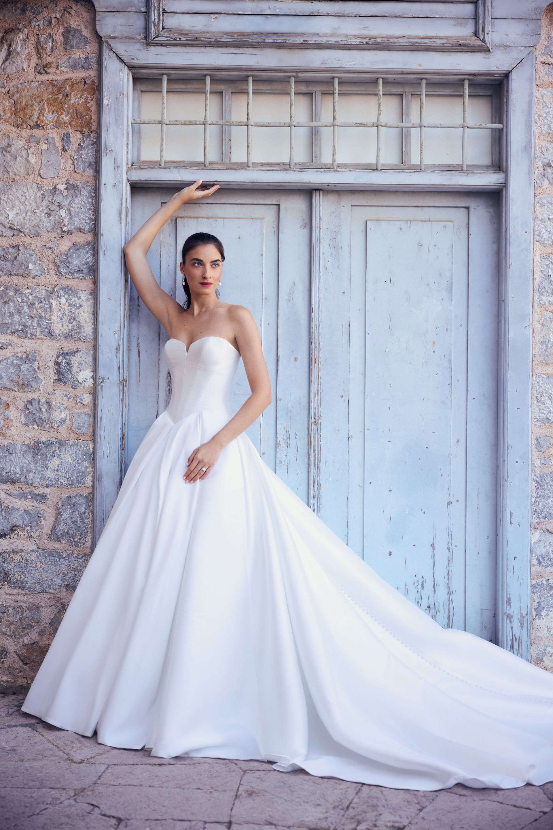 Woman in a white strapless gown poses by a blue door, arm raised. Long train spills onto the ground.