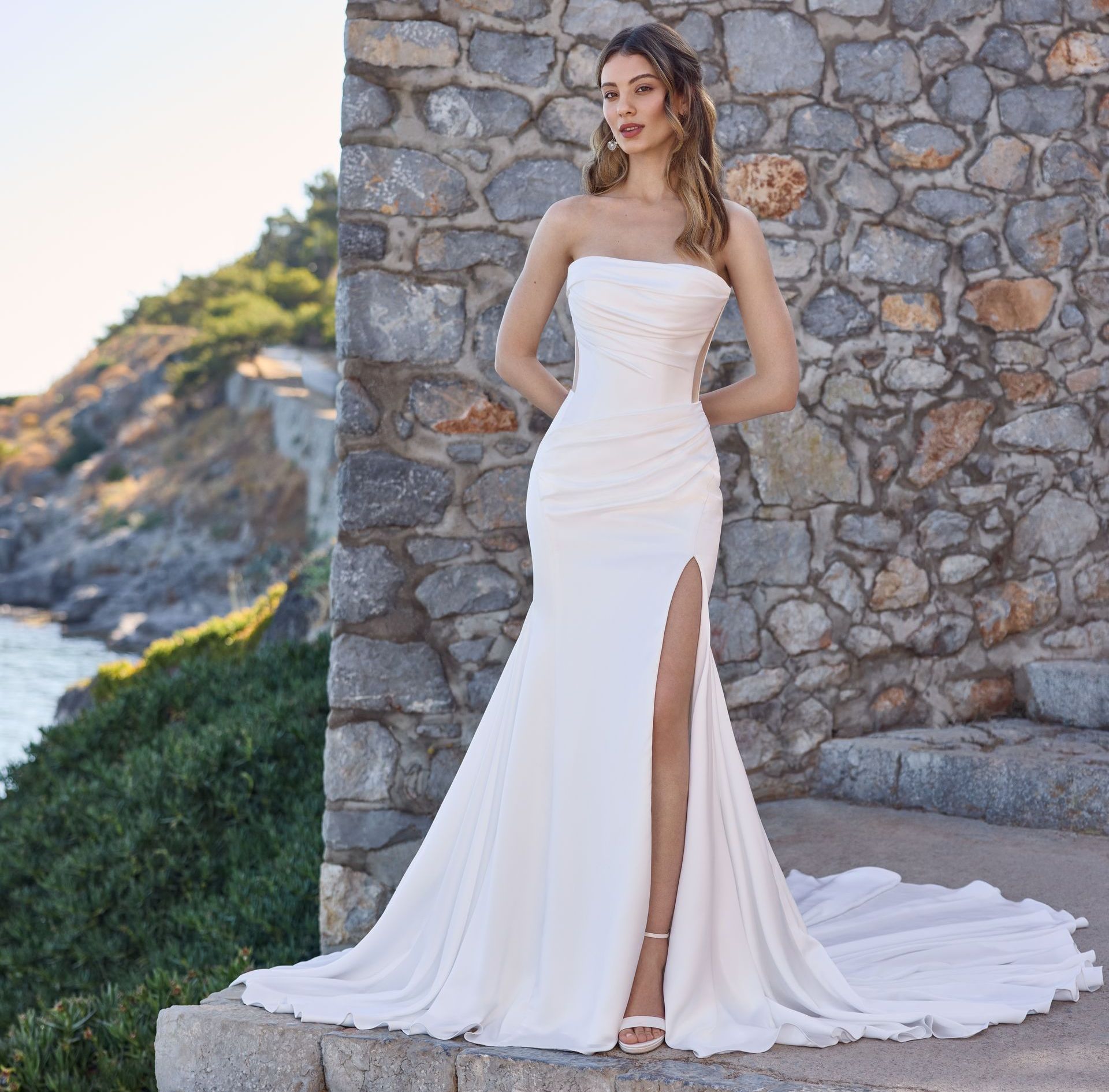 Woman in a white strapless gown with a high slit, standing on stone steps by a stone wall overlooking the ocean.