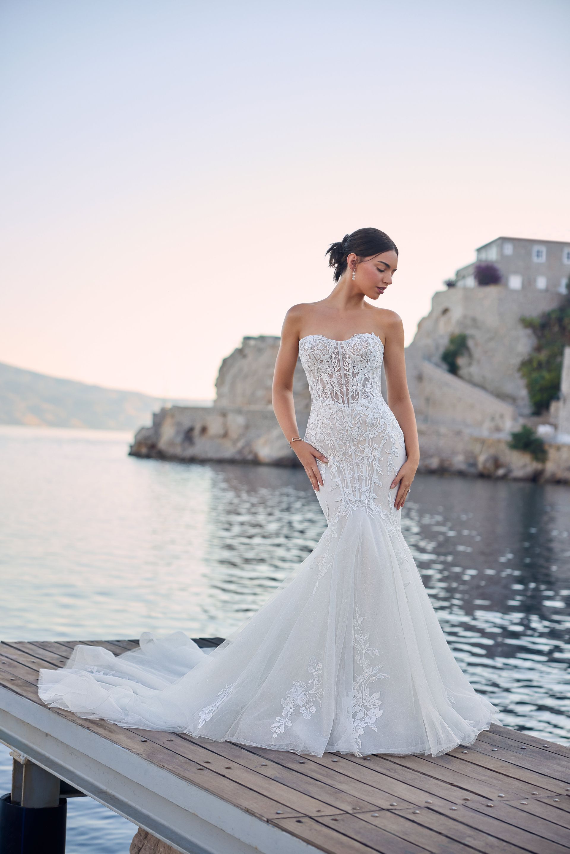 Woman in a strapless wedding dress poses on a pier overlooking the sea, near a rocky cliff.