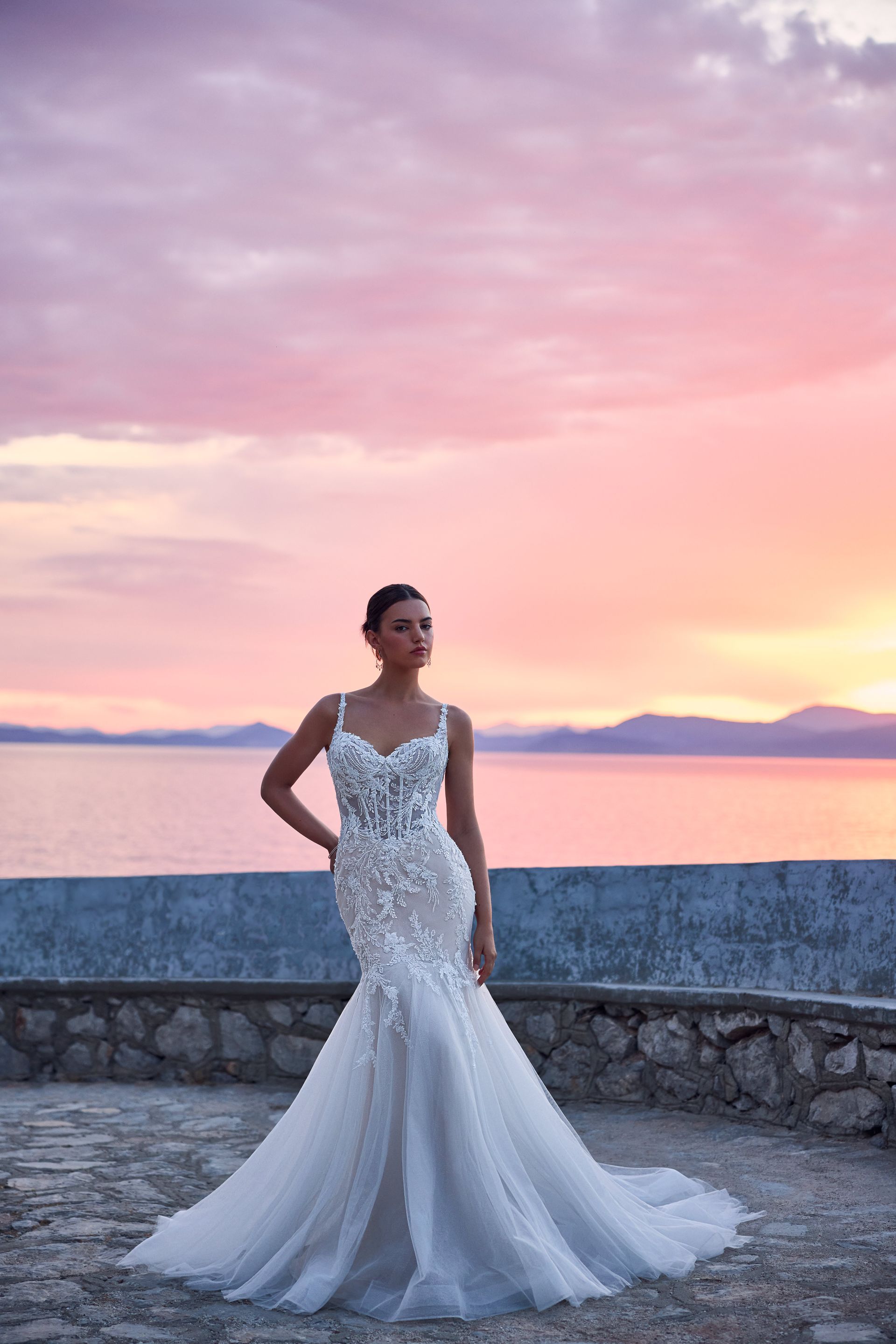 Woman in a white wedding dress stands at the edge of a sea. Sunset sky in the background.