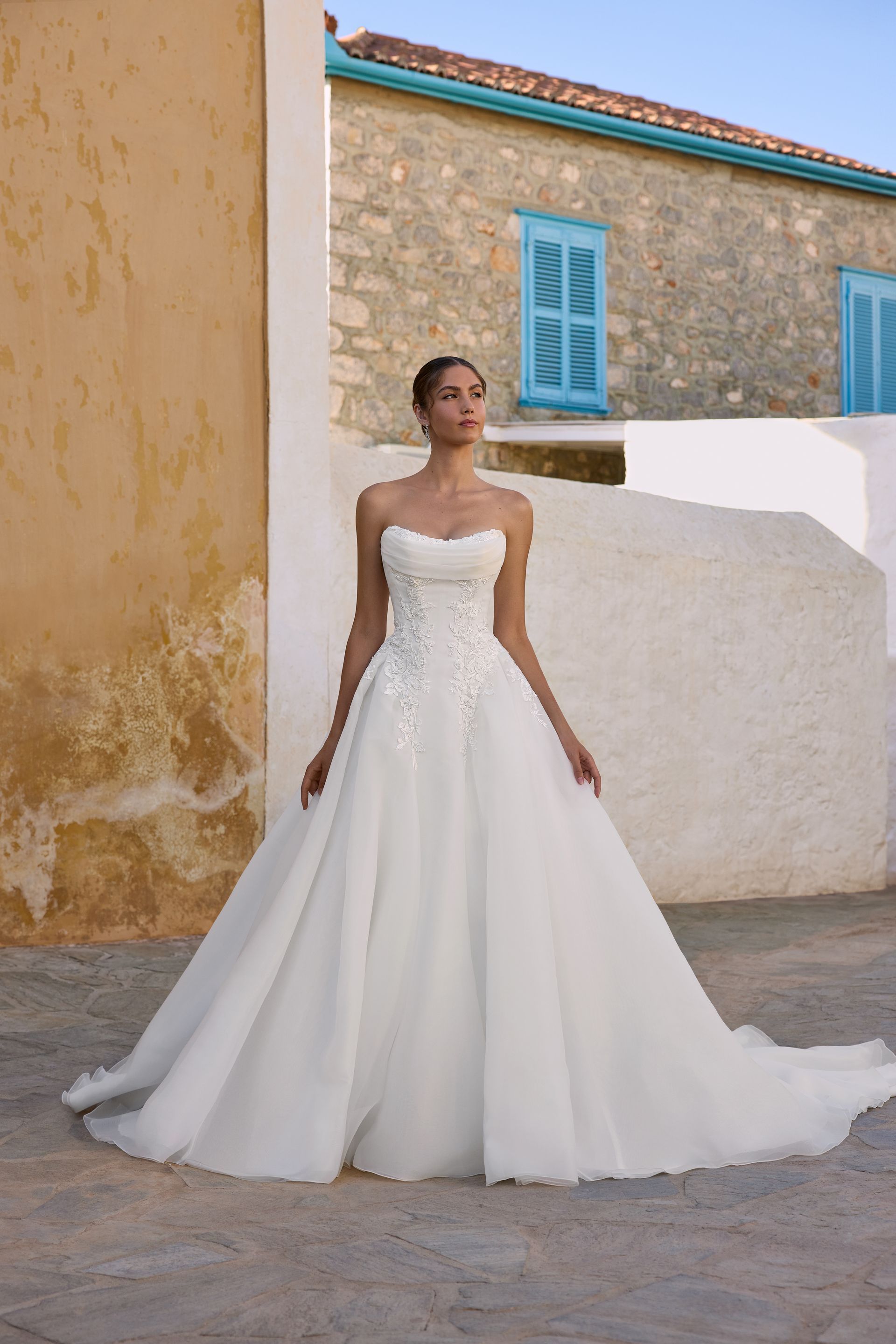 Woman in white strapless wedding gown outdoors, posing in front of a stone building with blue shutters.