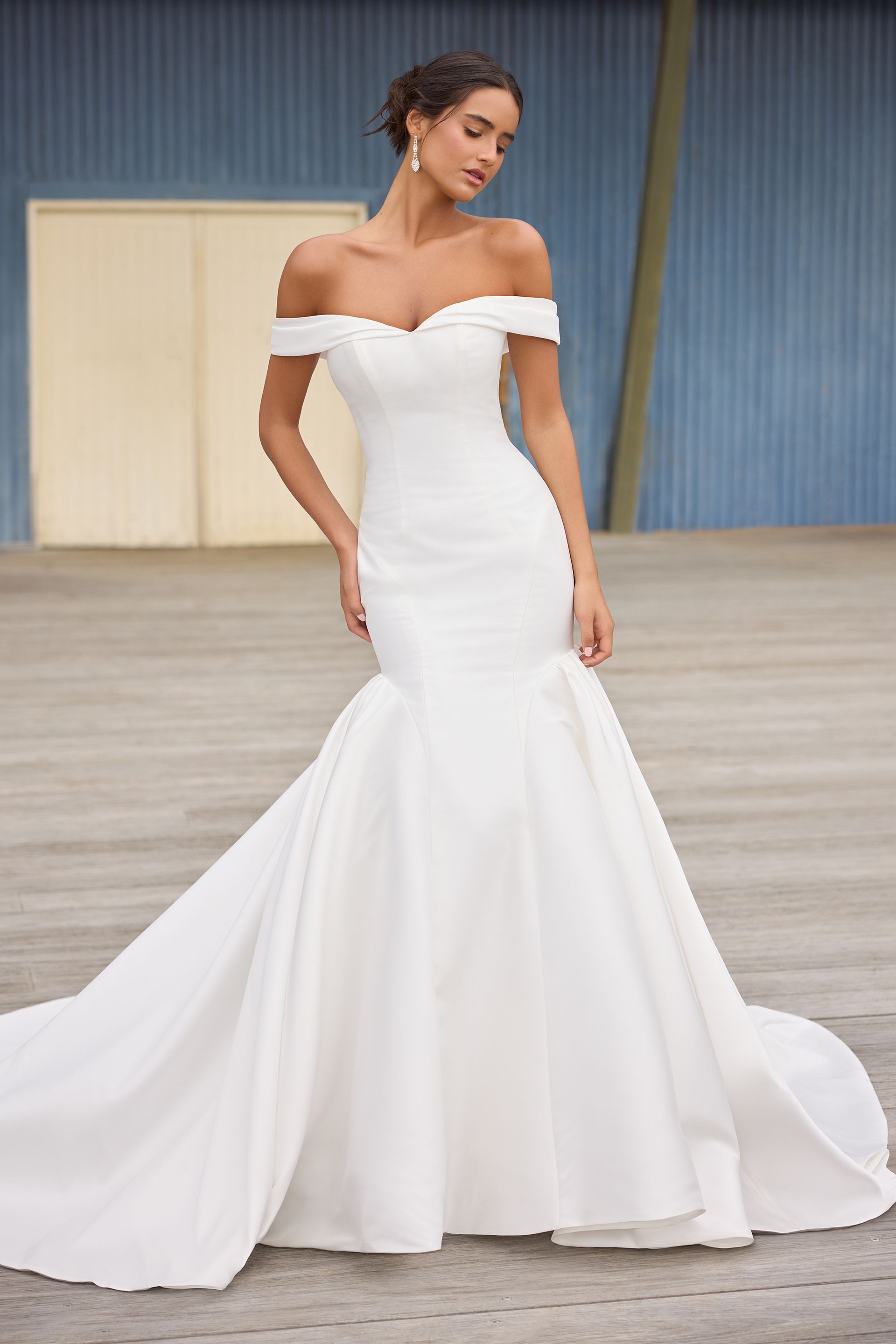 Woman in a white off-the-shoulder mermaid wedding gown, standing outdoors, looking down.