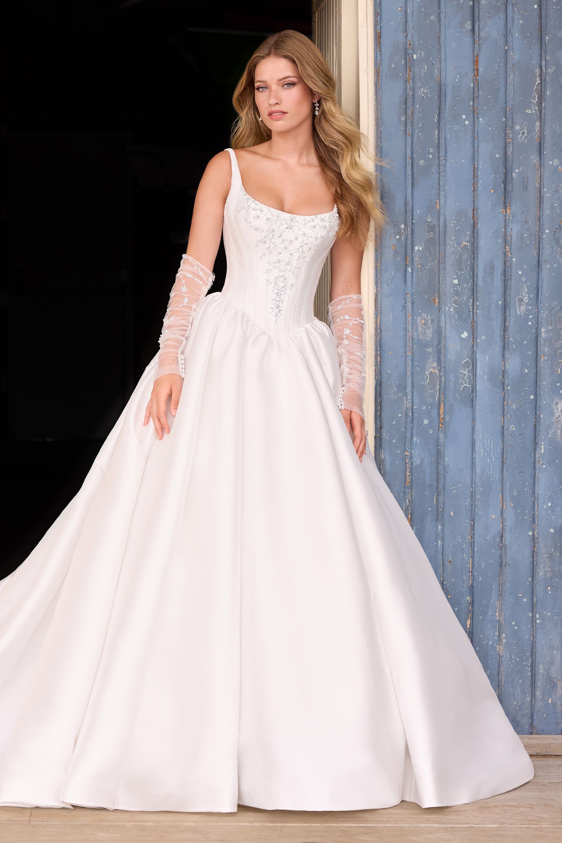 Woman in white wedding dress with lace gloves, standing in front of a blue wooden door.