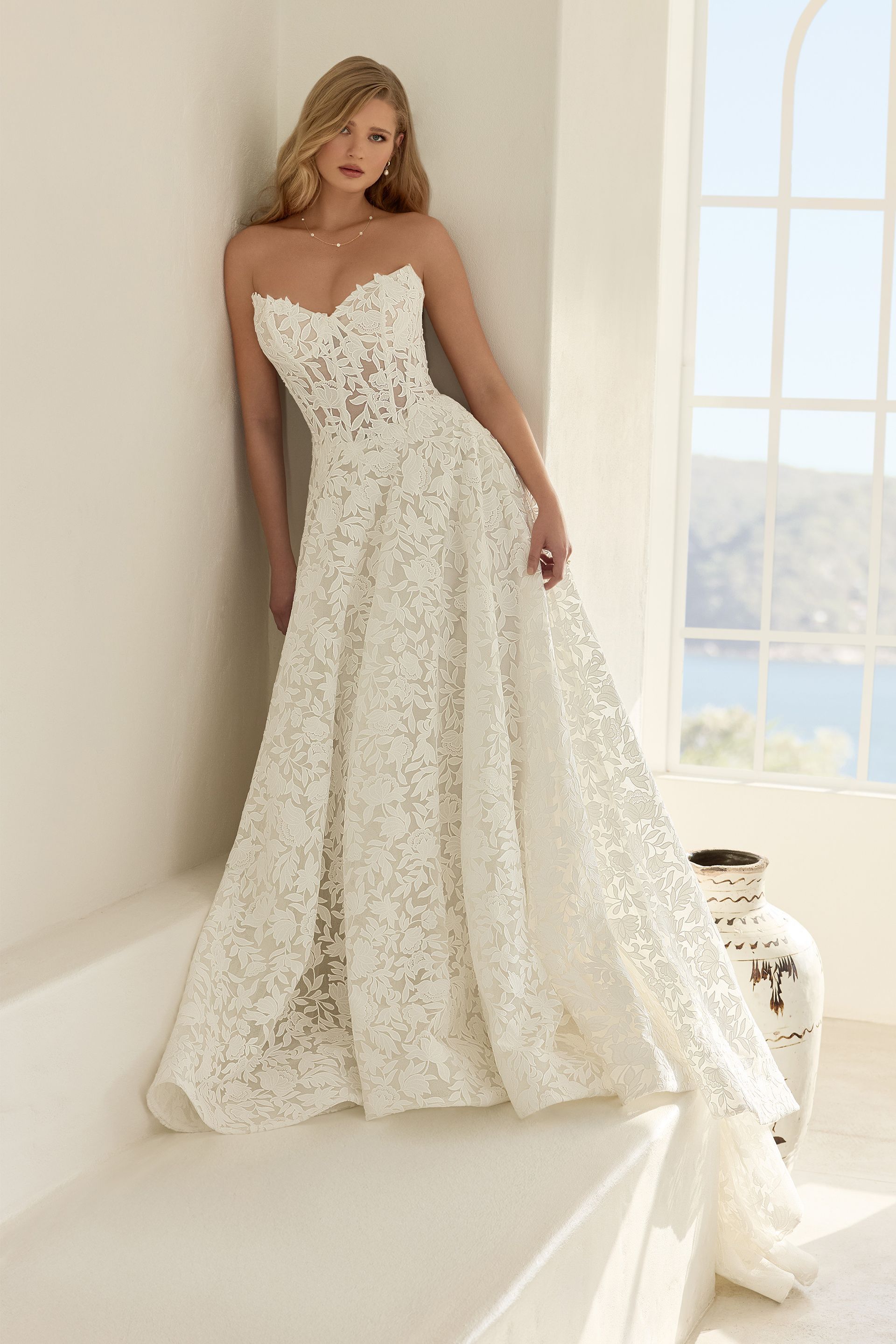 Bride in a strapless lace wedding gown standing by a bright window