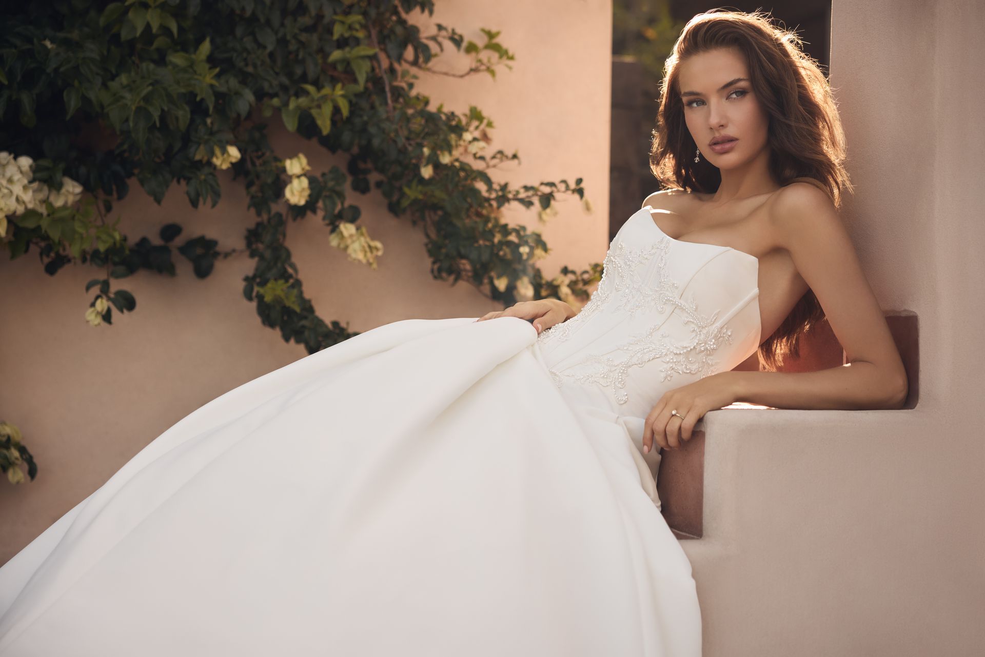 Woman in a white strapless dress reclining against a wall in a sunlit courtyard