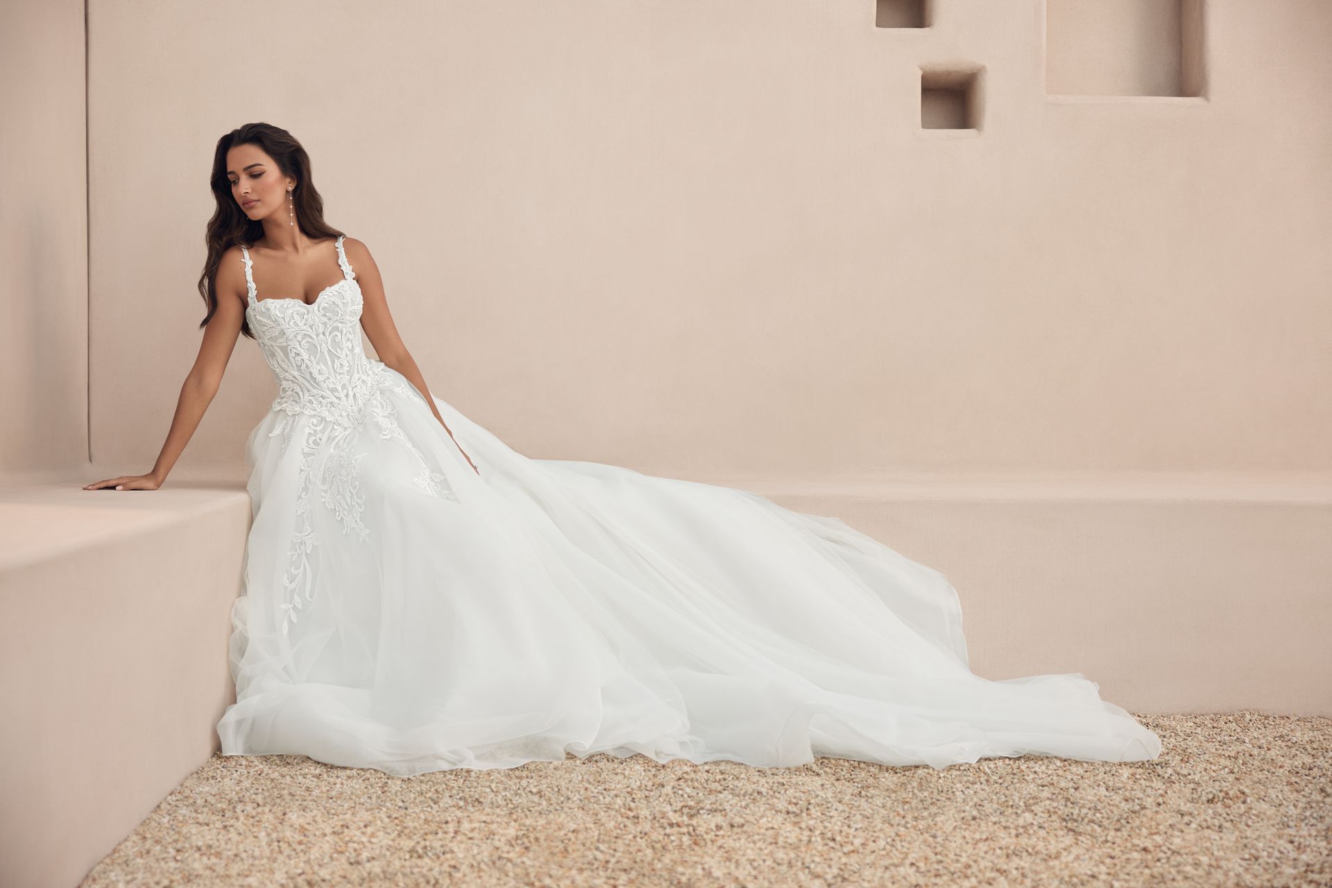 Woman in a white wedding gown posing against a beige wall with a long flowing train.