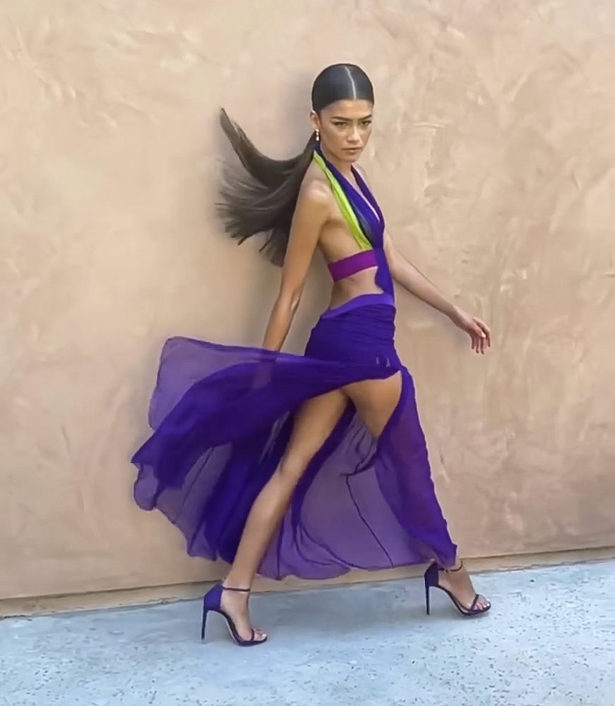 Woman in purple dress with side slit, walking pose, high heels, against beige wall.