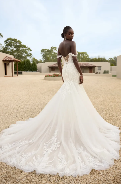 Bride in a white lace wedding gown with a long train, standing outdoors on a gravel courtyard.