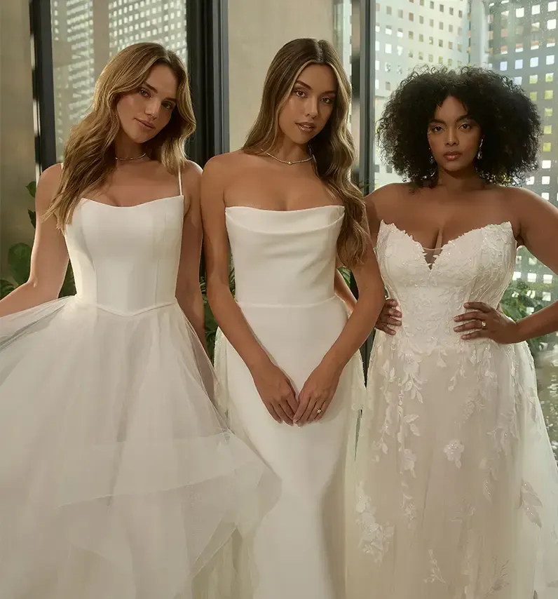 Three women in white wedding dresses standing together in a bright indoor setting. Three women in white wedding dresses standing together in a bright indoor setting.