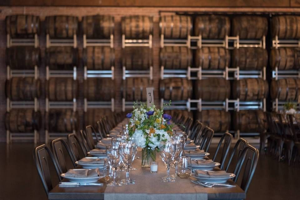 Long table set for a meal, with floral arrangement, in a winery with barrels on the wall.