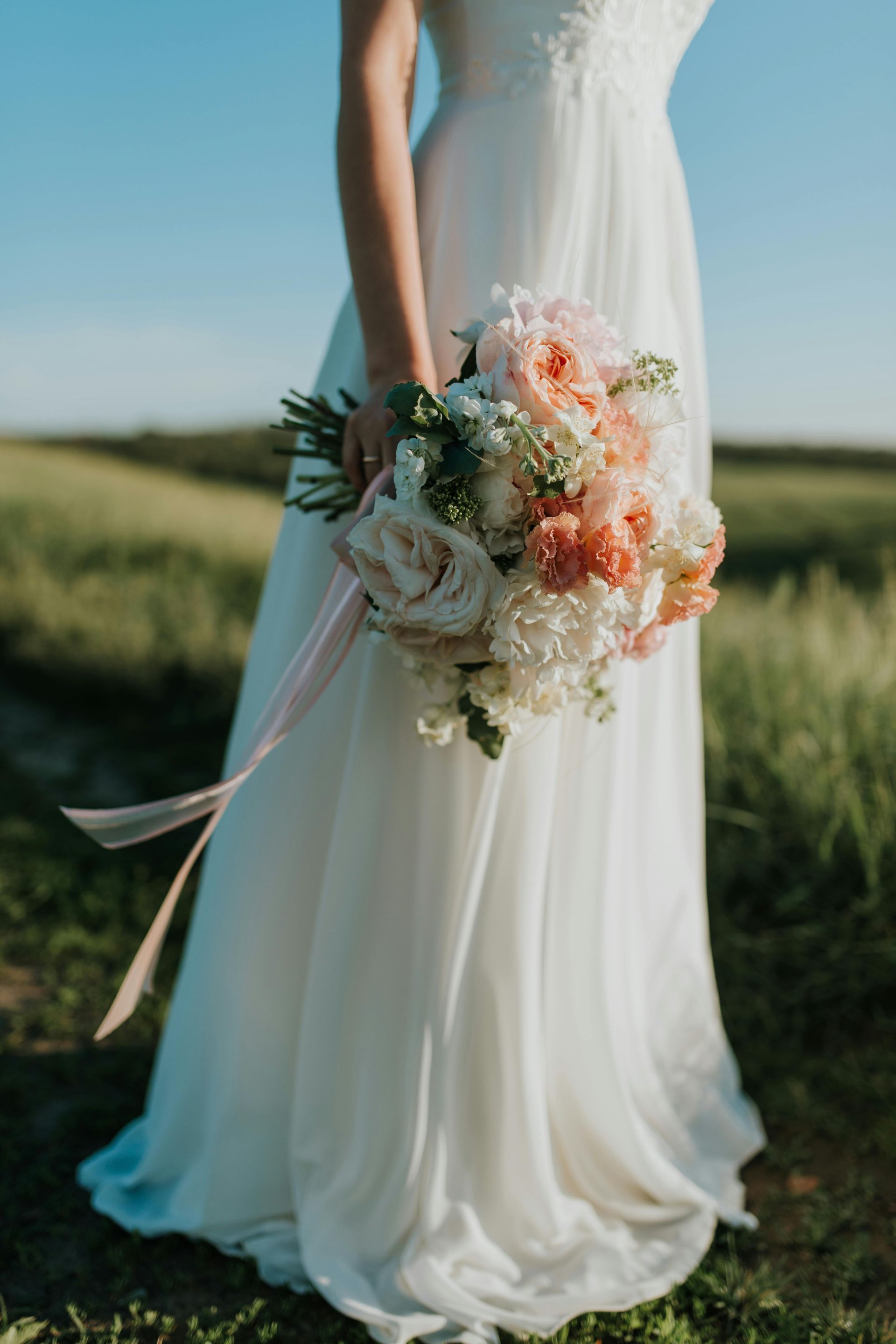 Bride in white dress holding a bouquet of peach and white flowers in a field of green.
