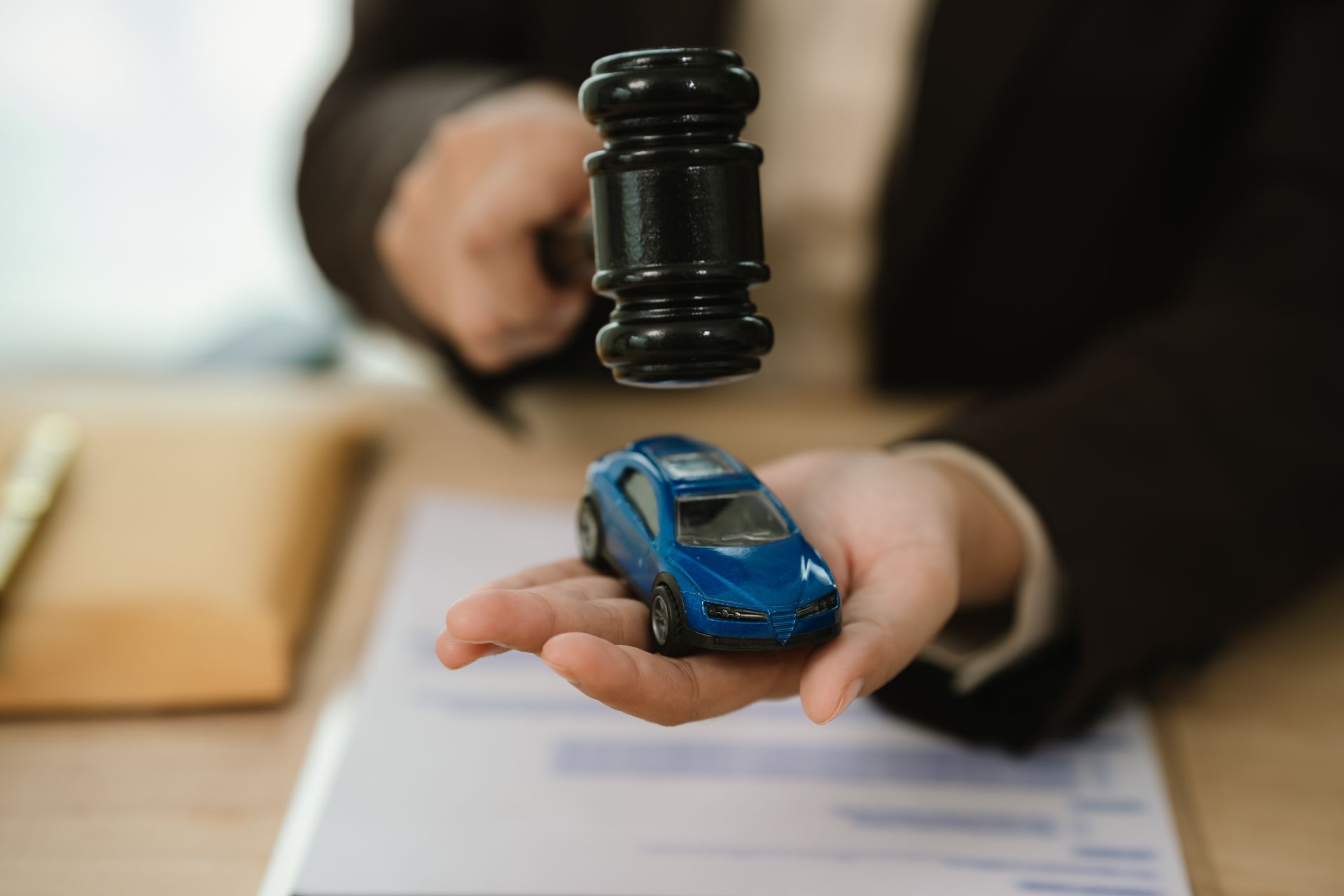 Hand holding a blue toy car while a judge’s gavel hovers above it on a desk