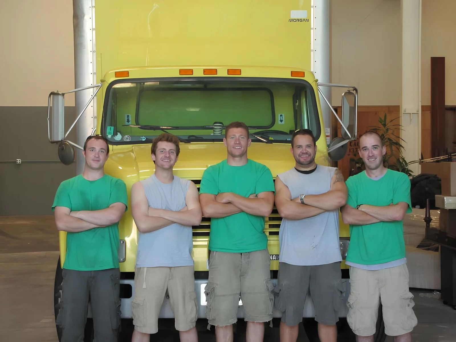 Five people in green shirts and gray sleeveless tops stand with arms crossed in front of a yellow truck in a warehouse.