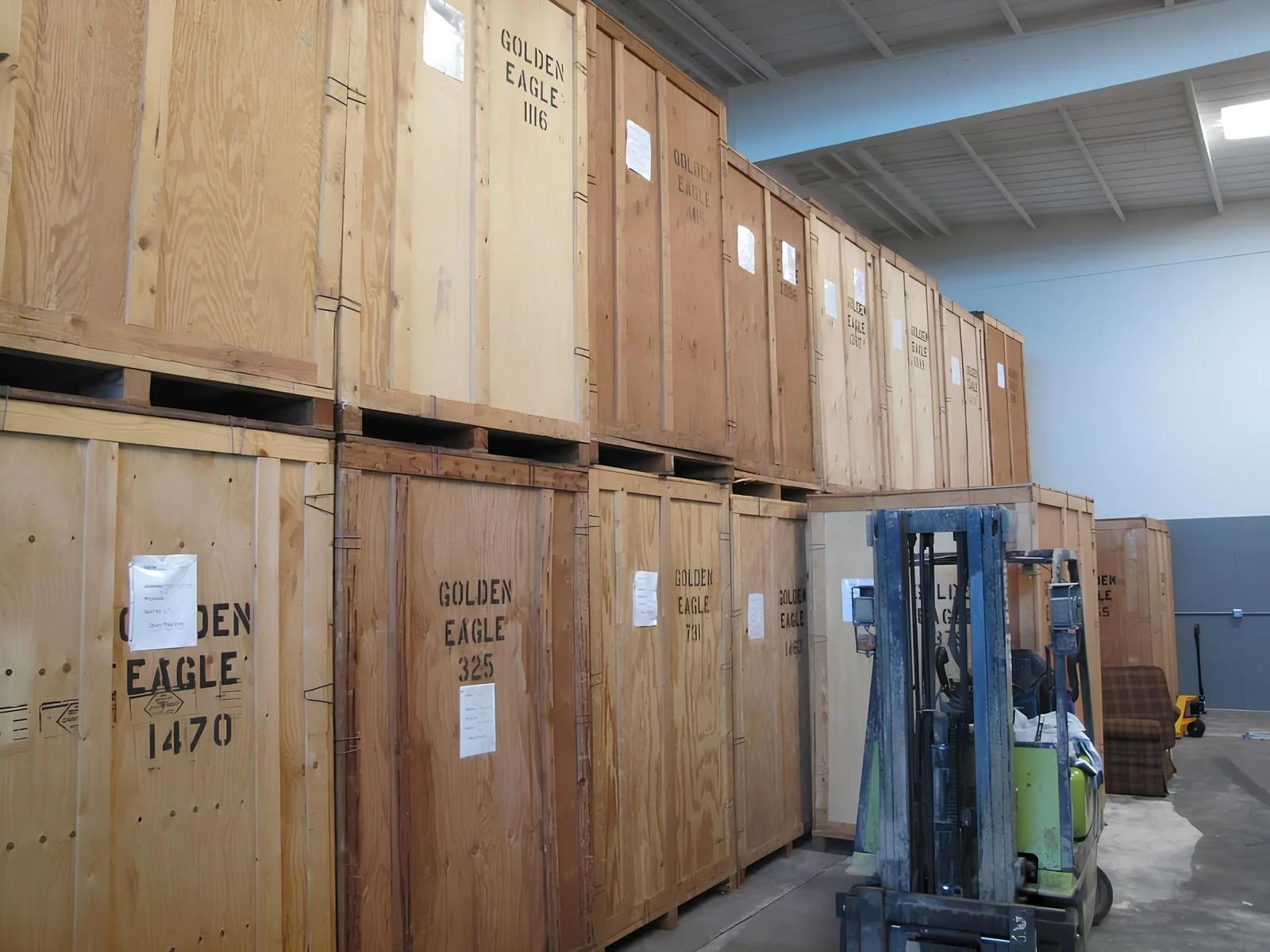 A warehouse interior showing wooden storage crates stacked two levels high with a forklift parked in the aisle.