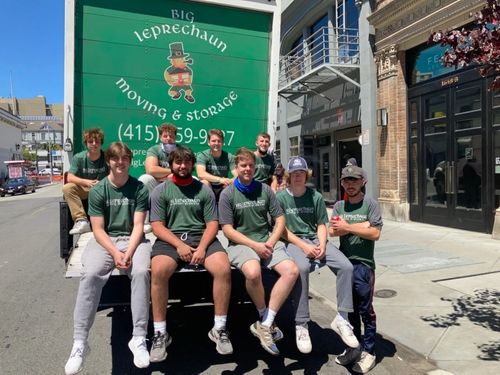 A group of nine people in green uniform shirts posing on the back of a Big Leprechaun Moving & Storage truck.