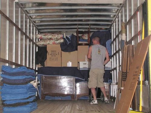 A person in a gray tank top and beige shorts moves boxes inside a truck filled with furniture and packing blankets.