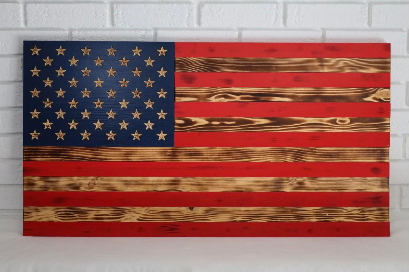 Wooden American flag with blue stars and red and wood grain stripes against a white brick wall.