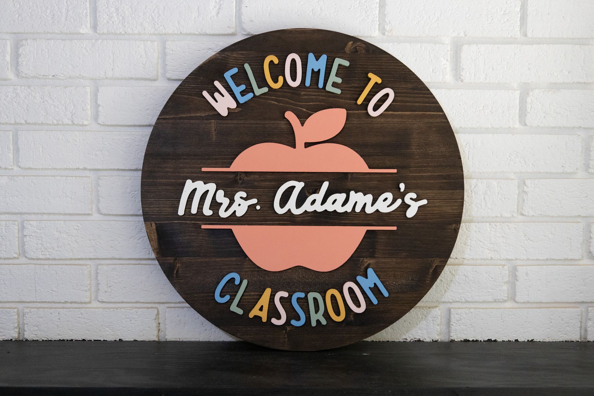 Round classroom sign reading “Welcome to Mrs. Adams’s Classroom” with an apple on a white brick wall