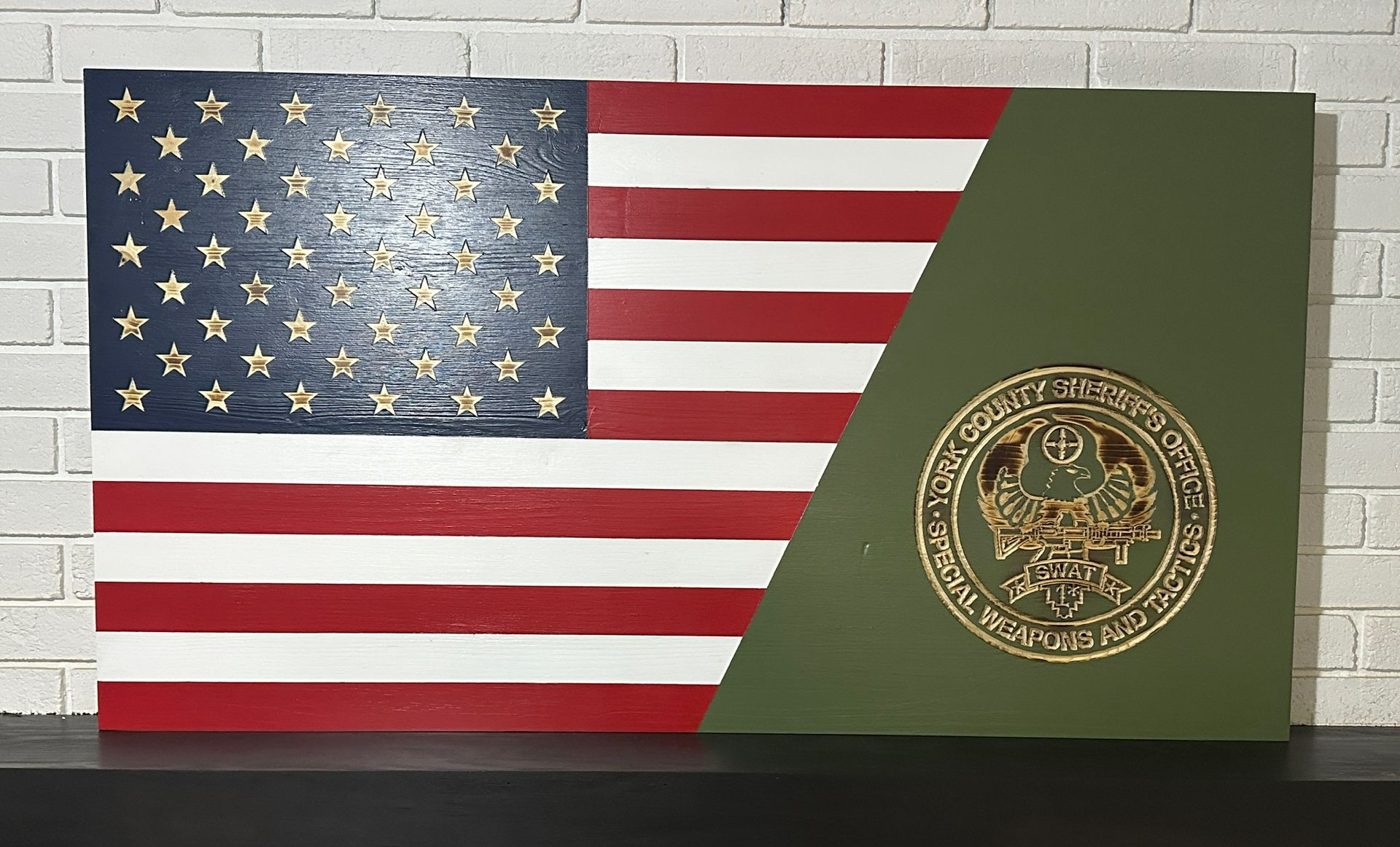 Wooden artwork: American flag with green section, gold seal, against a white brick wall.