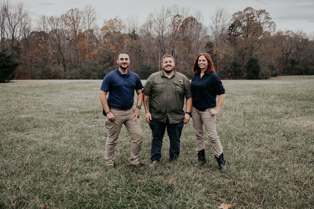 Three people standing in a field. Trees in the background. Man in center wearing olive shirt smiles. Others in blue shirts.